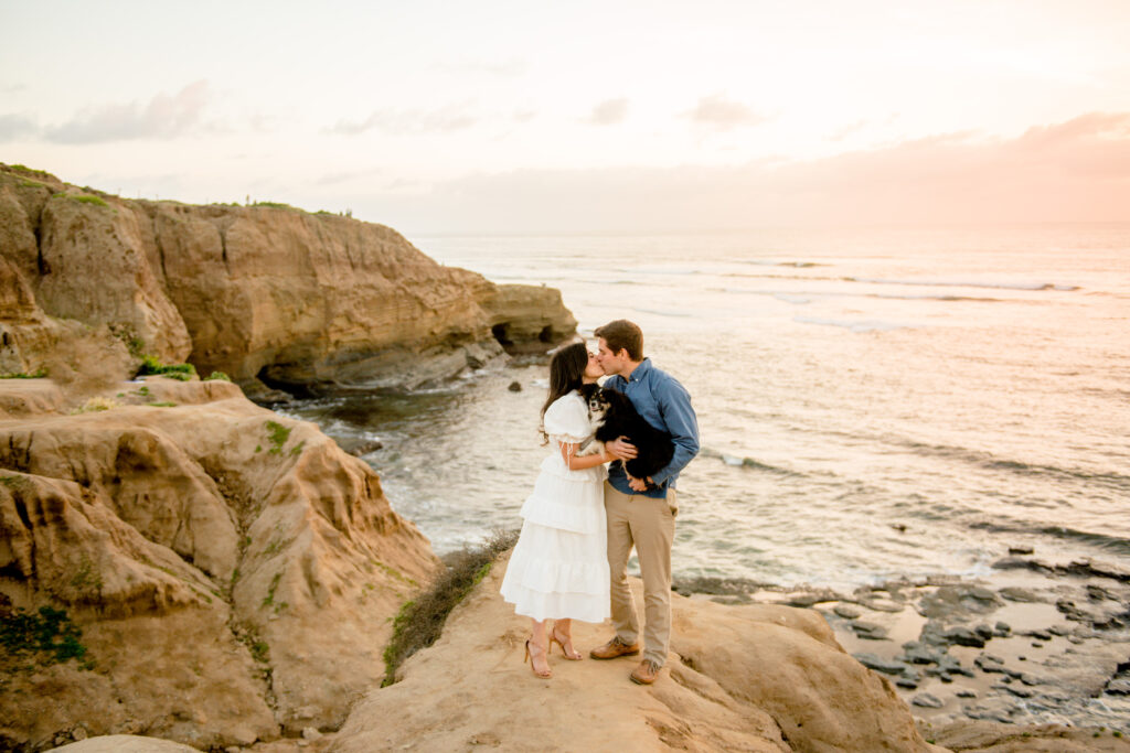 beautiful couple sharing a romantic kiss on a cliff overlooking the ocean. Their dog is snuggled up between them looking at the camera. In the background is a beautiful pink sunset. 