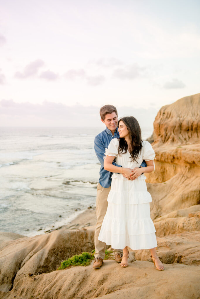 couple is standing on the cliffside overlooking the ocean in san diego. The woman is staring at the sun while her partner is smiling at her. 