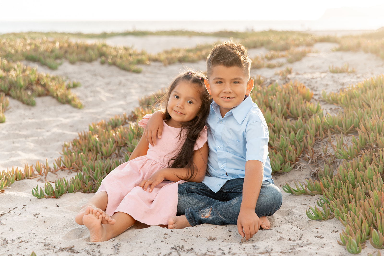A young boy in a blue shirt and jeans sits on a beach with an arm around his younger sister at sunset