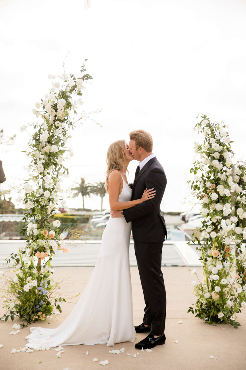 NEwlyweds kiss under a flower arch of awhite roses in a black tuxedo and white silk dress at a l'auberge del mar wedding