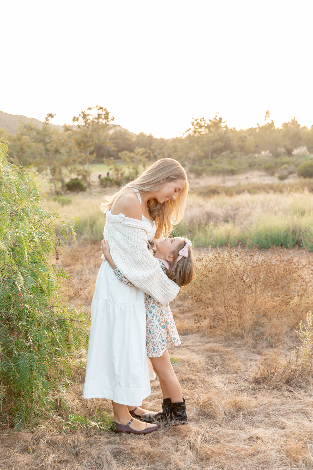 A mom and daughter hug while standing in a field at sunset