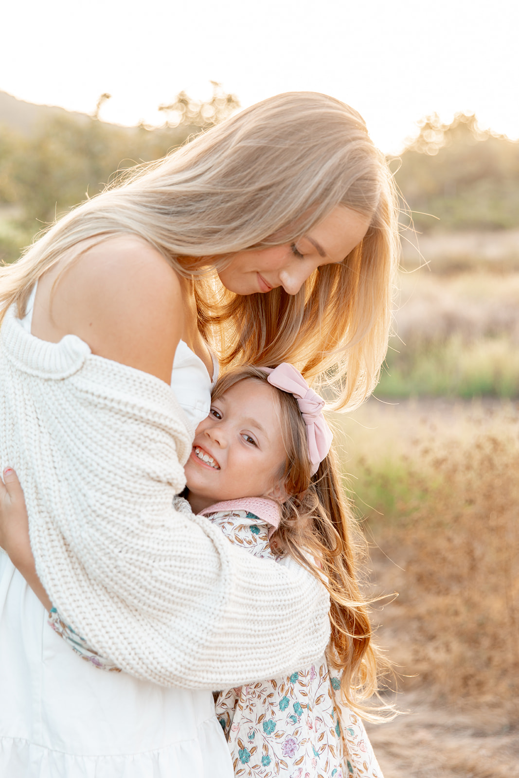 A mother in a white knit sweater bends over hugging her young daughter in a field at sunset after attending mommy and me classes in san diego