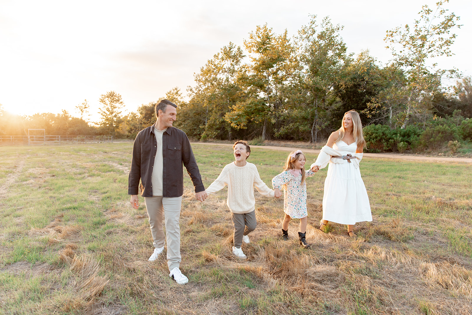 A mom and dad walk through a grassy field holding hands with their smiling son and daughter at sunset after mommy and me classes in san diego