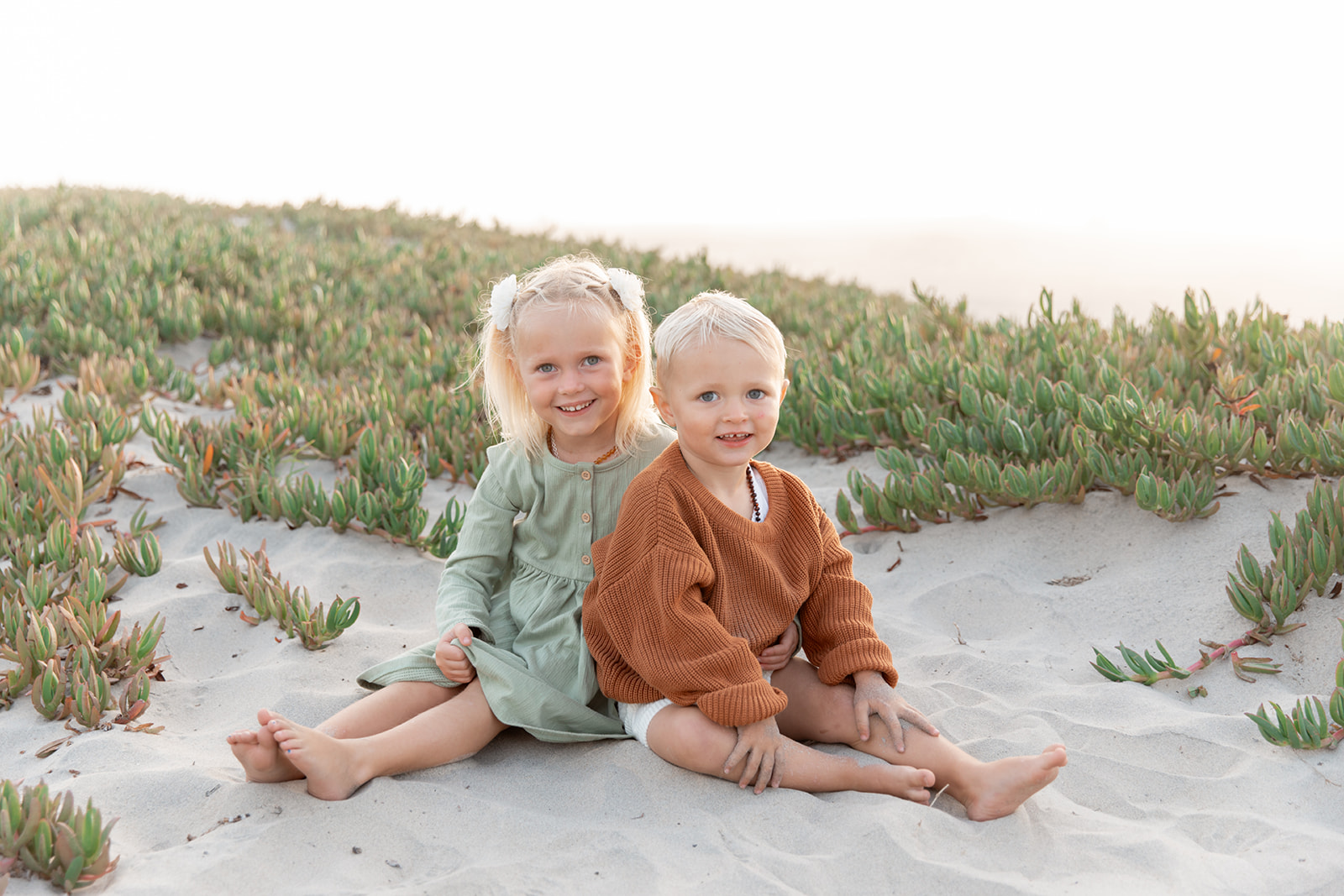 A young girl and her younger brother sit on a beach dune at sunset hugging while wearing a green dress and orange sweater
