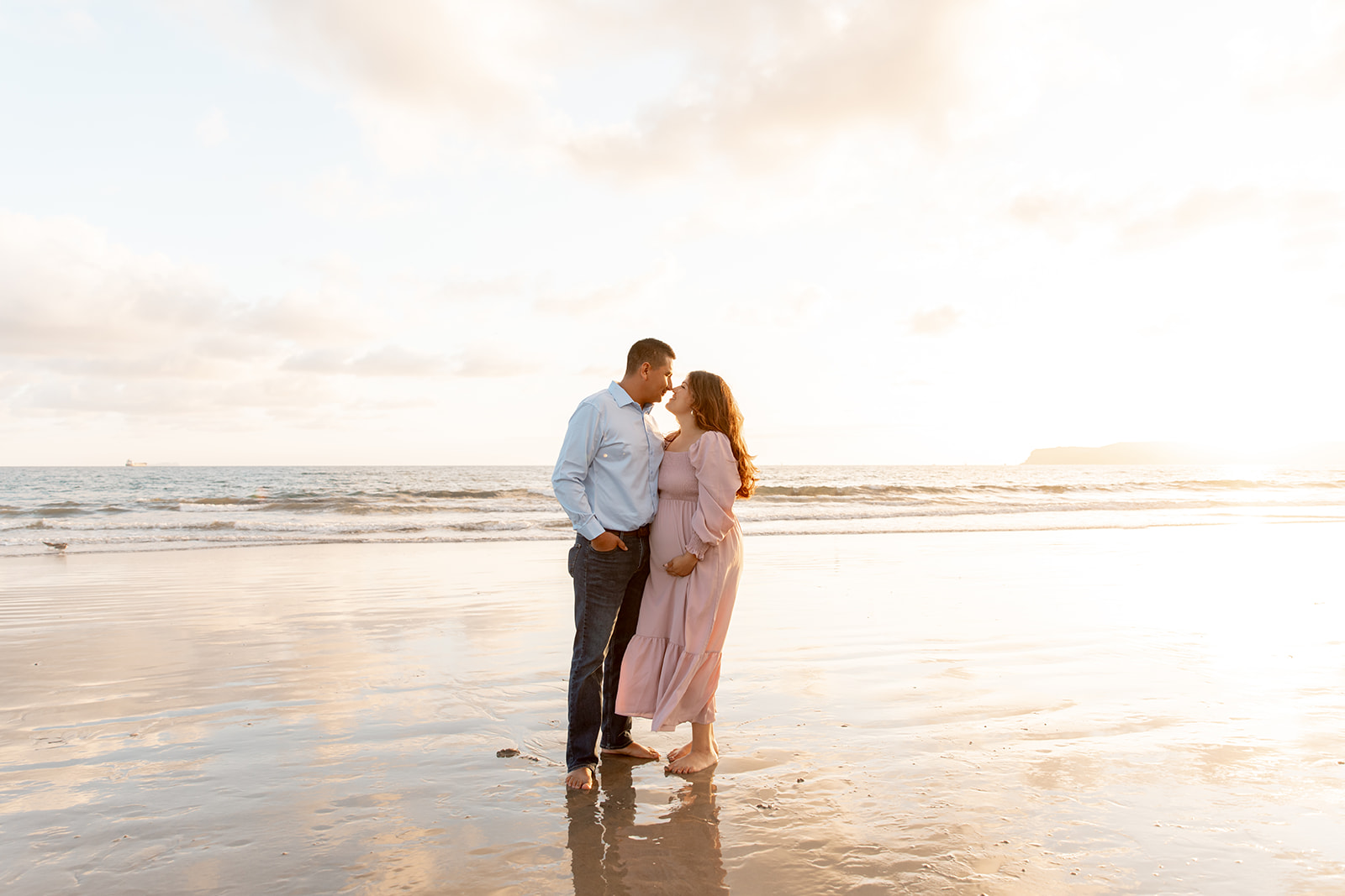 A mom to be stands in a pink maternity dress kissing her husband in a blue shirt on a beach at sunset