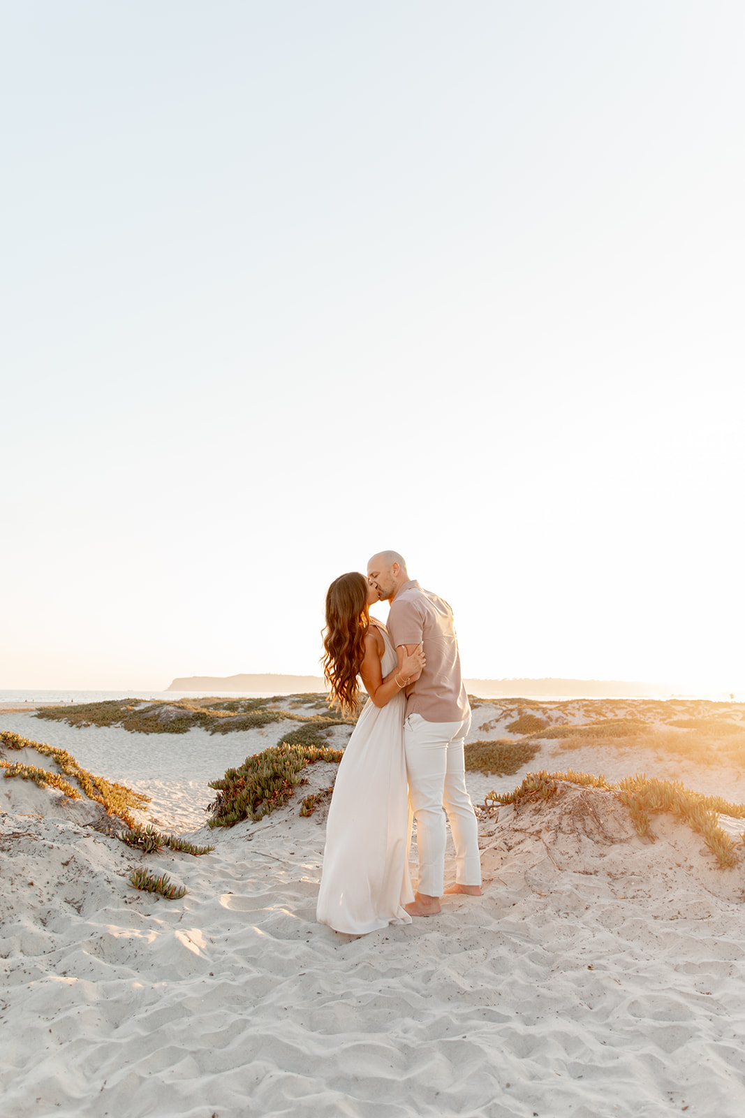 A husband and wife kiss while standing in the dunes of a beach at sunset while their kids visit San Diego Daycares