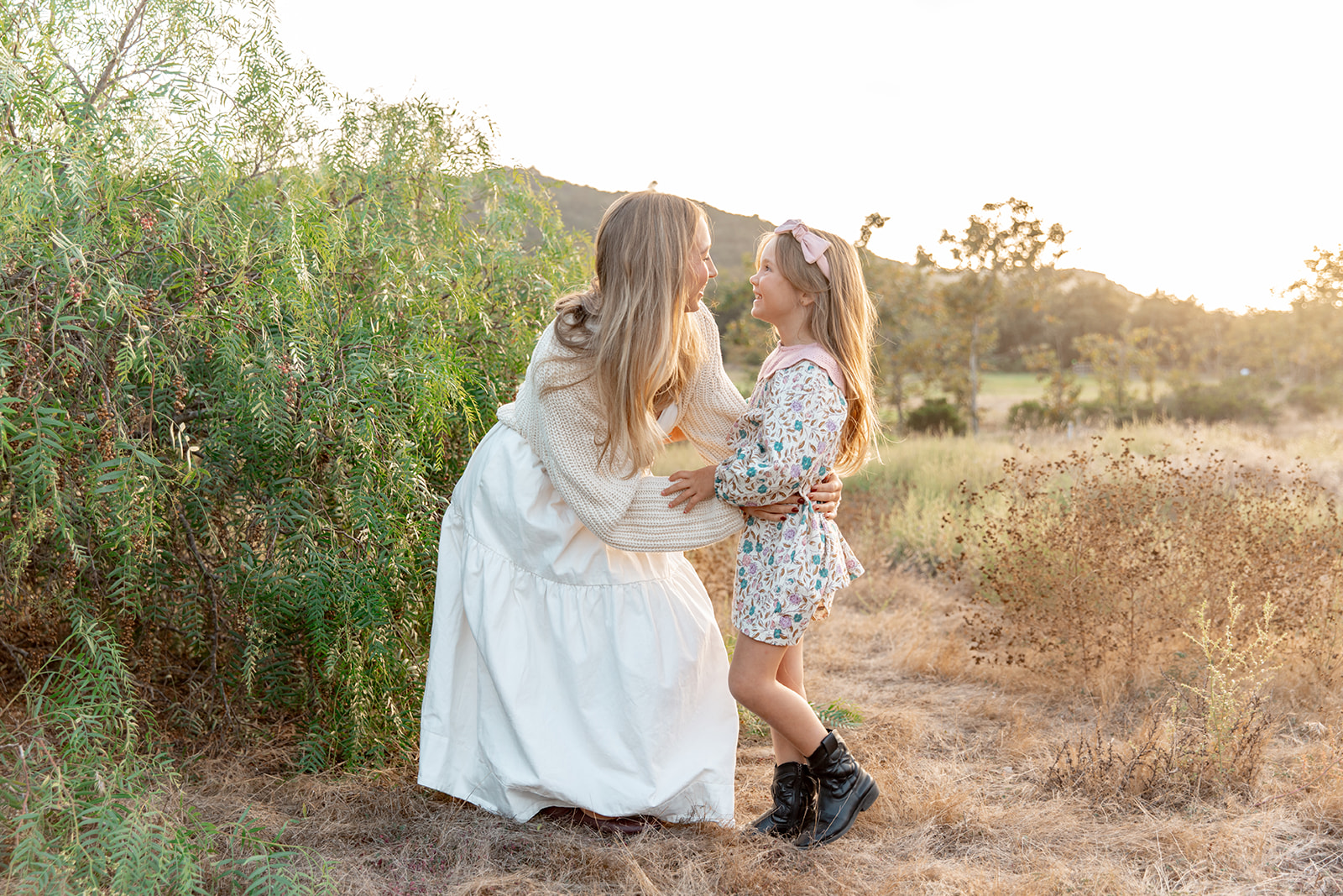 A mother in a white dress hugs onto her her daughter in a colorful floral dress while standing in a grassy park at sunset San Diego Family Restaurants