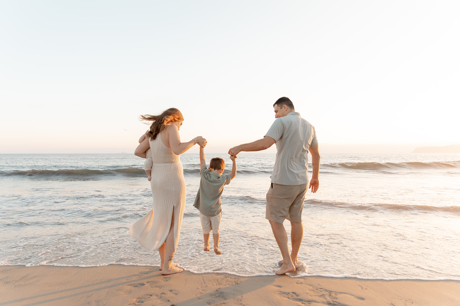 A mom and dad hold the hands and lift their toddler son above a wave on the beach at sunset