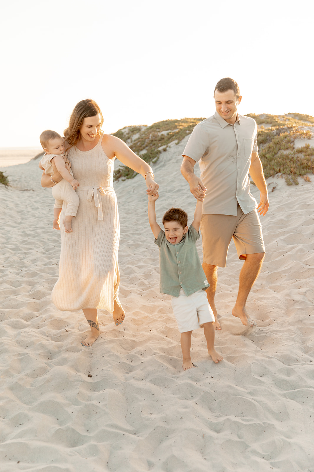 A mom and dad swing and play with their toddler son while walking up a beach while mom holds their infant on her hip San Diego Pediatric Dentists