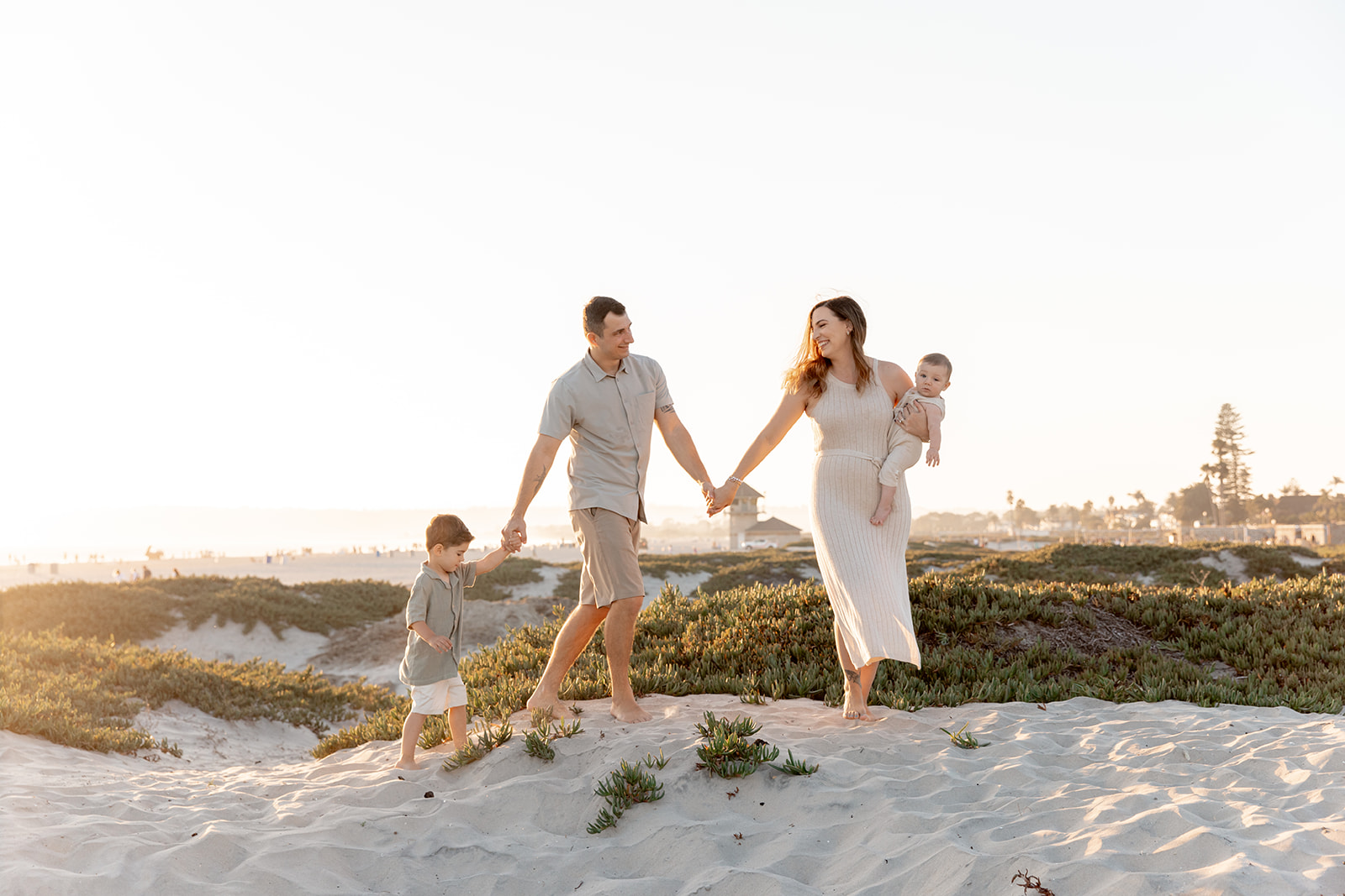 A mother and father walk holding hands up a beach dune with their toddler son and mom holding their infant on her hip with big smiles from San Diego Pediatric Dentists