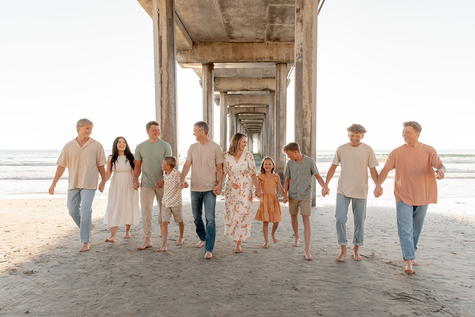 A large family of ten walk up a beach under a pier holding hands and looking and smiling at each other San Diego Pediatricians