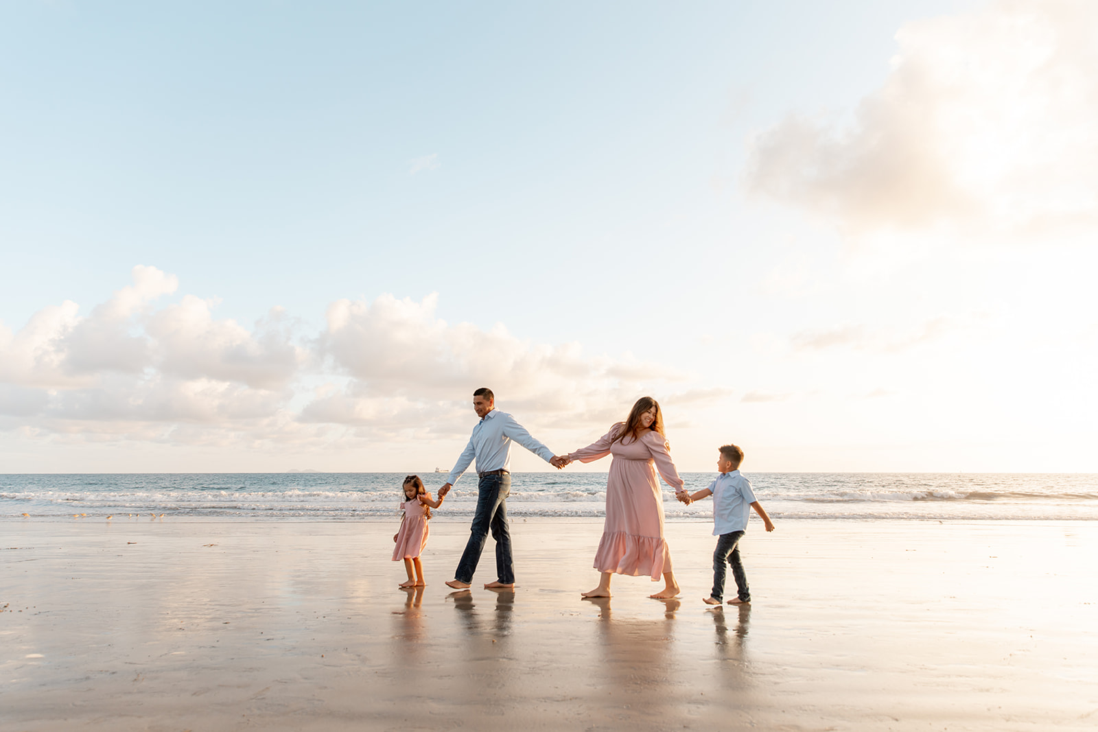 A mom and dad in a pink dress and blue shirt walk hand in hand with their young daughter and son in matching outfits to theirs down a beach at sunset after visiting San Diego Pediatricians
