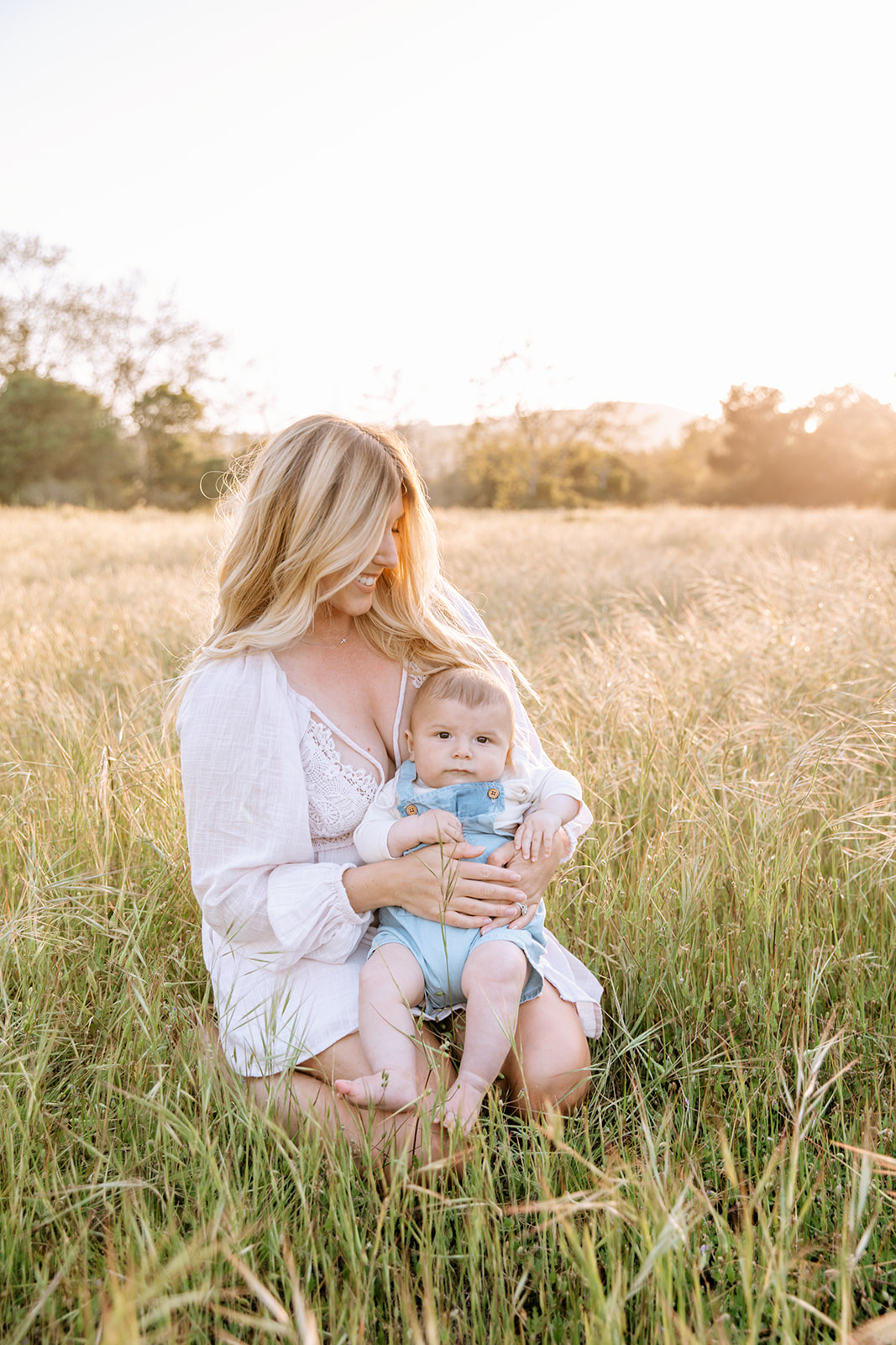 A new mom kneels in a field of tall grass with her infant son in her lap wearing blue overalls at sunset after a visit to The New Children's Museum