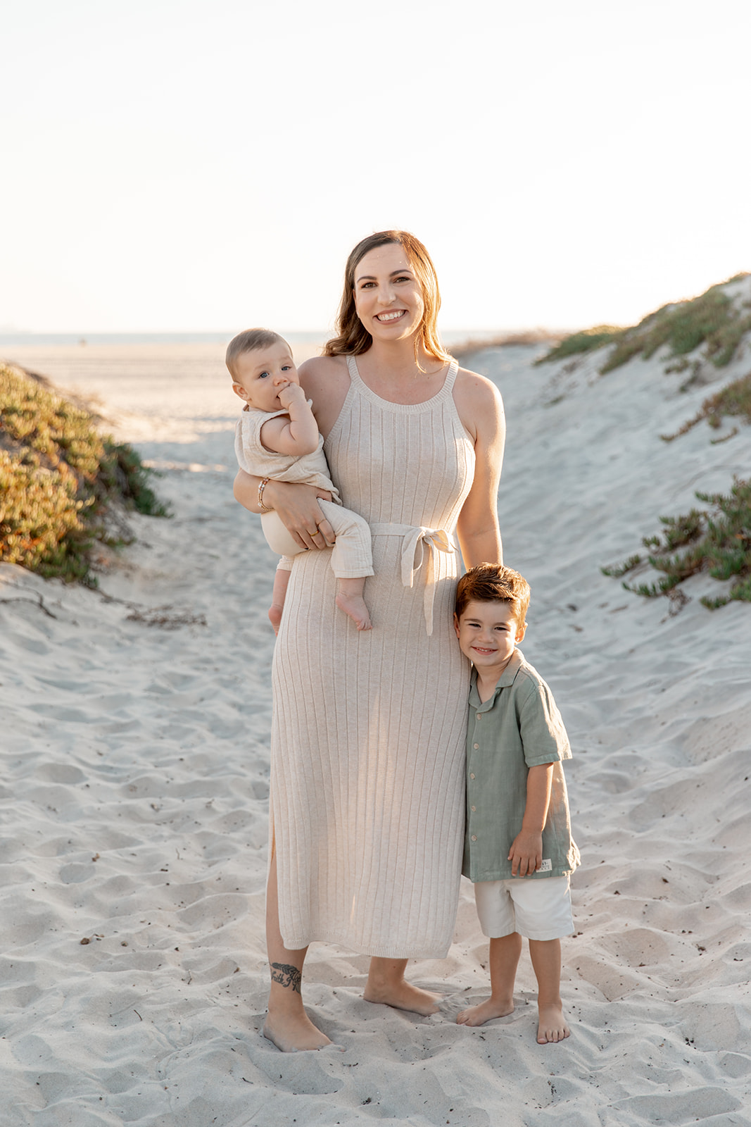 A mom stands in a beach path with her toddler son at her side and infant on her hip at sunset