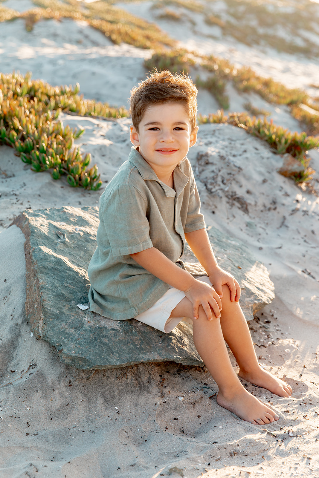 A young boy sits on a rock at the beach at sunset in a green button down shirt while doing toddler activities in san diego