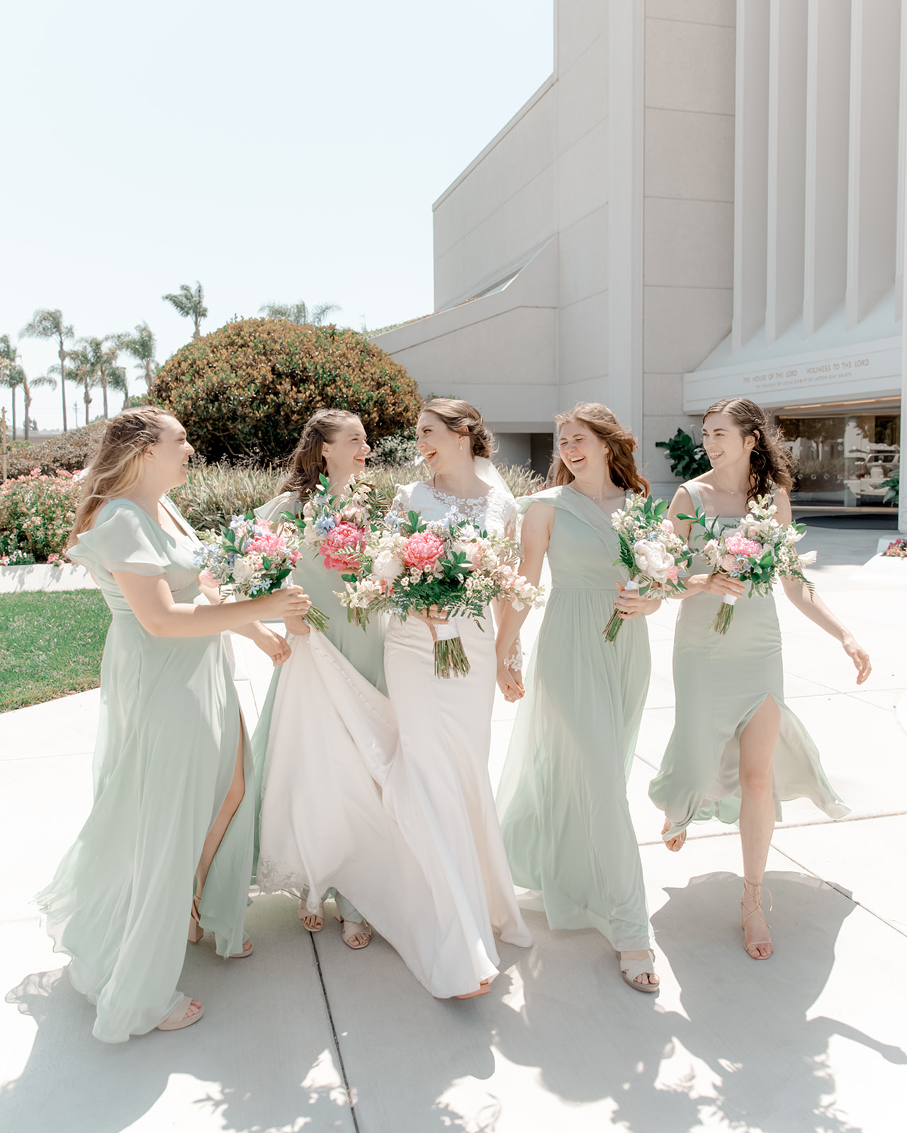A bride in a white silk dress walks through a park with her bridesmaids in sage dresses all holding white and pink bouquets