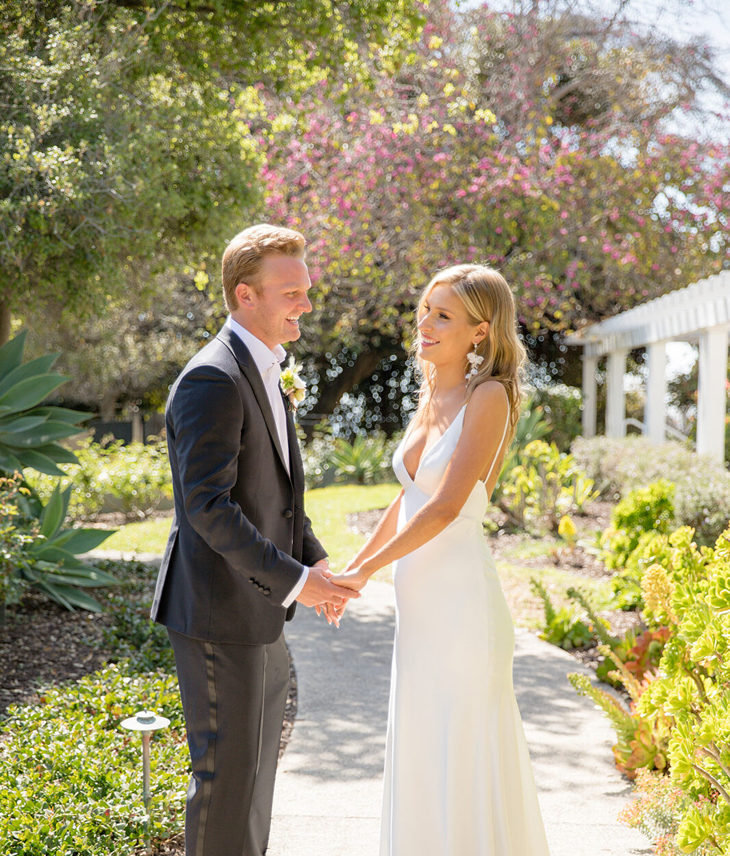 Newlyweds hold hands and stand in a garden path laughing at each other in a black tuxedo and white silk dress at a Vista Valley Country Club wedding