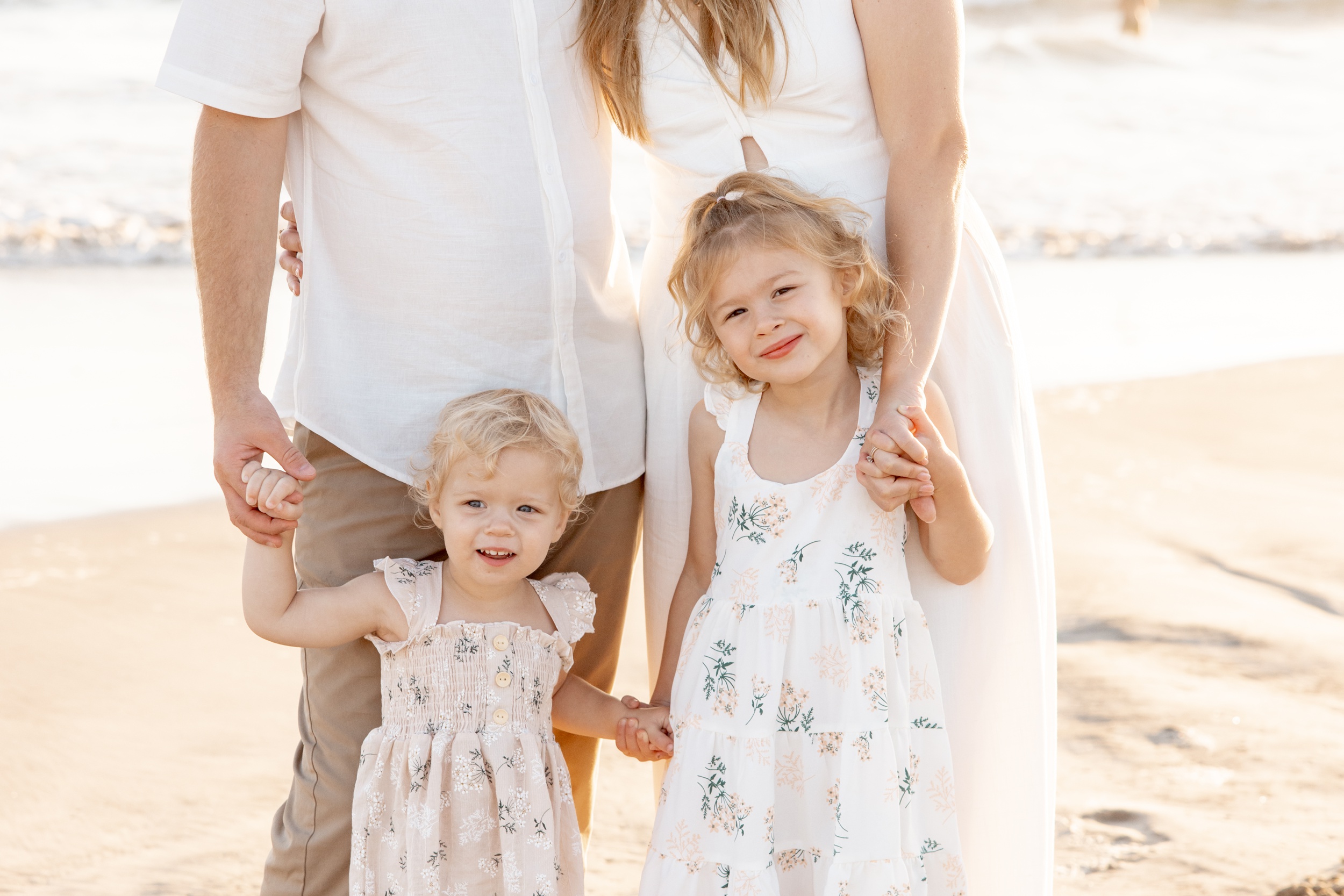 Two toddler sisters stand holding hands with each other and mom and dad in white dresses on a beach