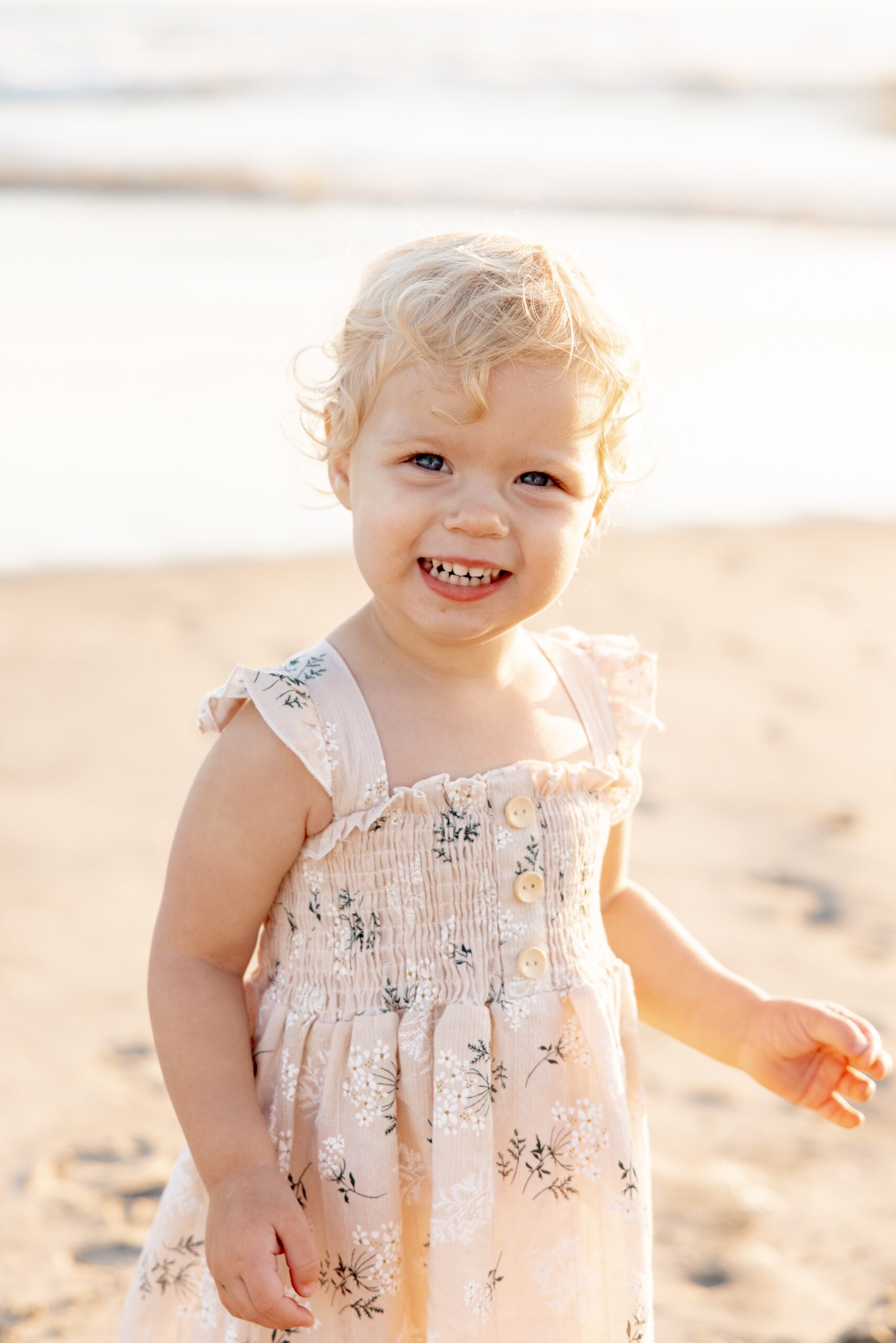 A giggling toddler in a pink dress plays in the sand at the beach after visiting children's boutiques in san diego