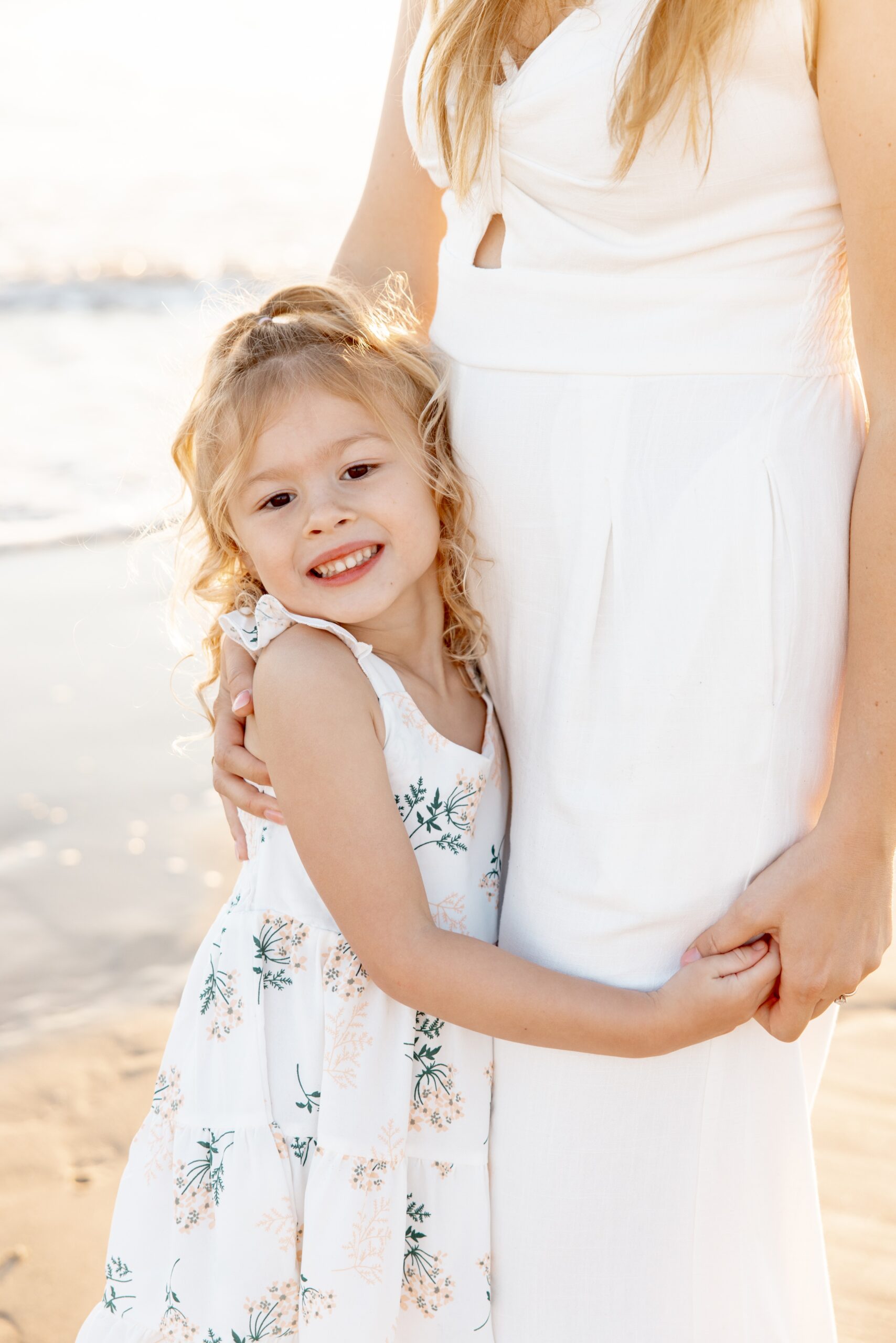 A smiling young girl in a white flower dress hugs her mom in a white gown on a beach at sunset after visiting children's boutiques in san diego