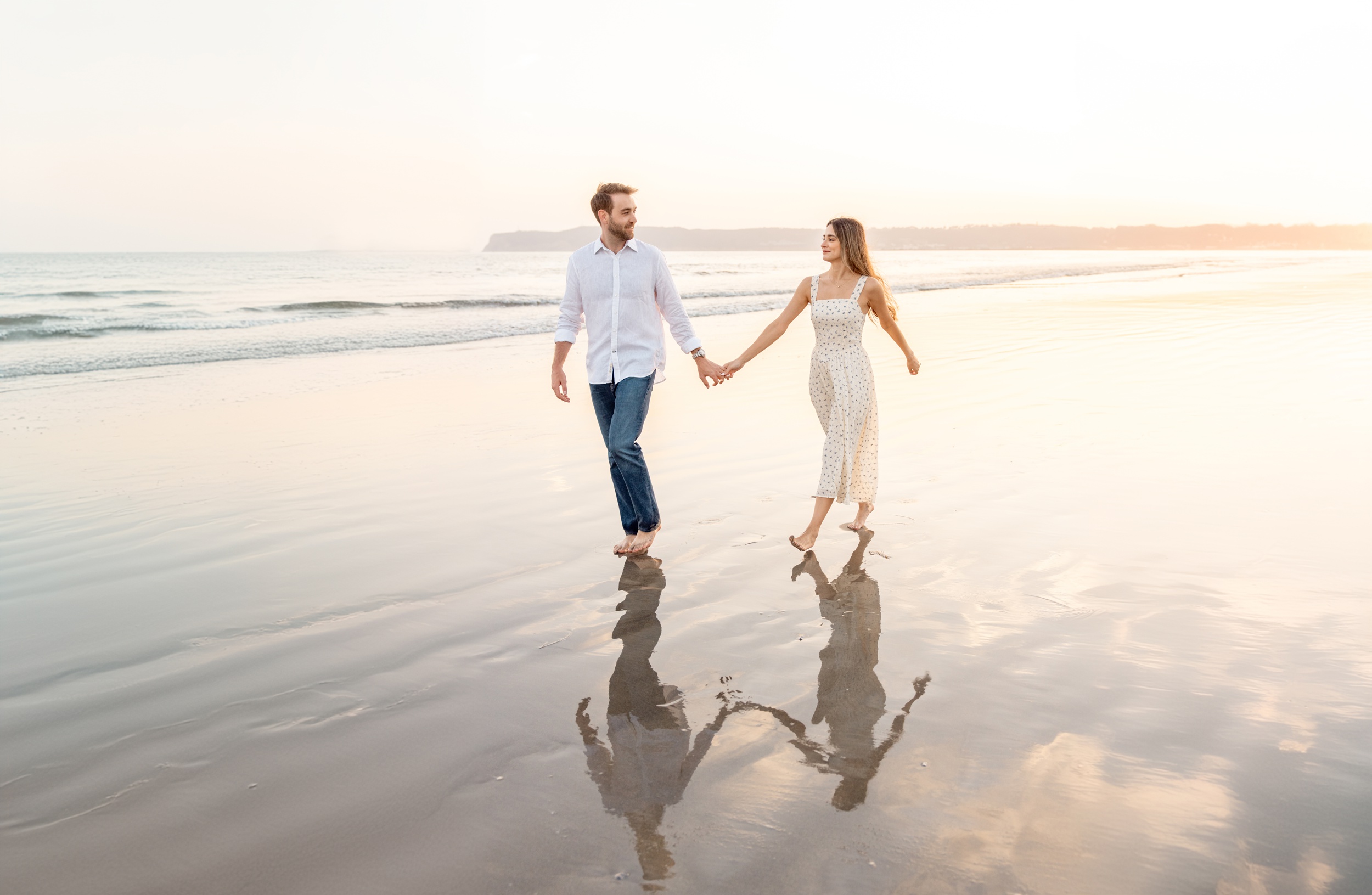 A happy couple walks on a beach in white at sunset holding hands before eloping in san diego