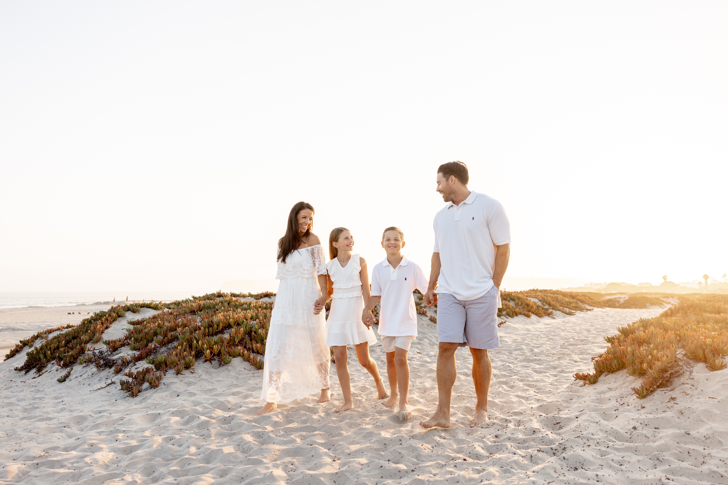 A happy family of 4 hold hands and walk on a beach at sunset dressed in white after some horseback riding lessons for kids san diego