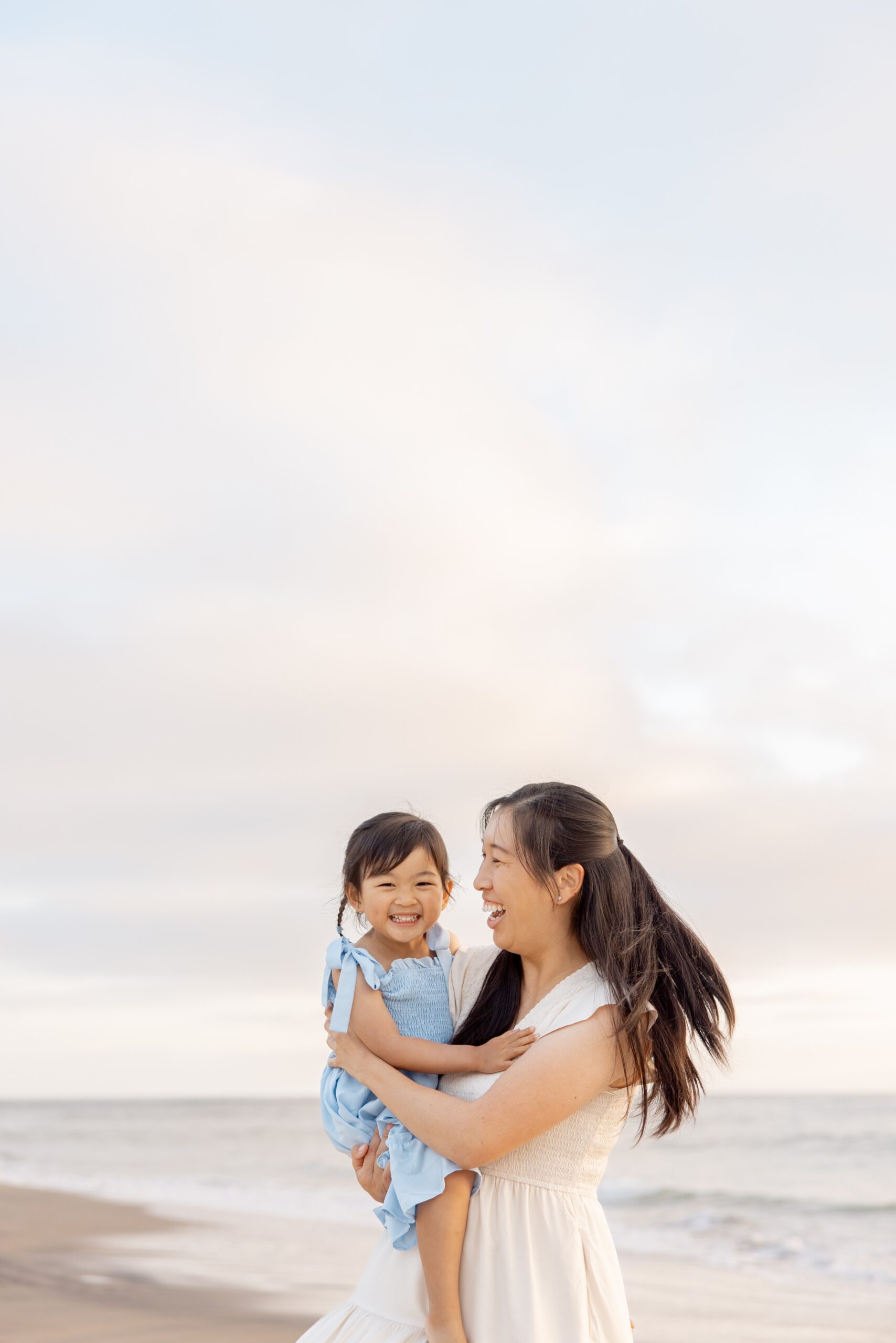 A giggling toddler girl dances with mom on a beach in a blue dress after finding a montessori daycare in san diego