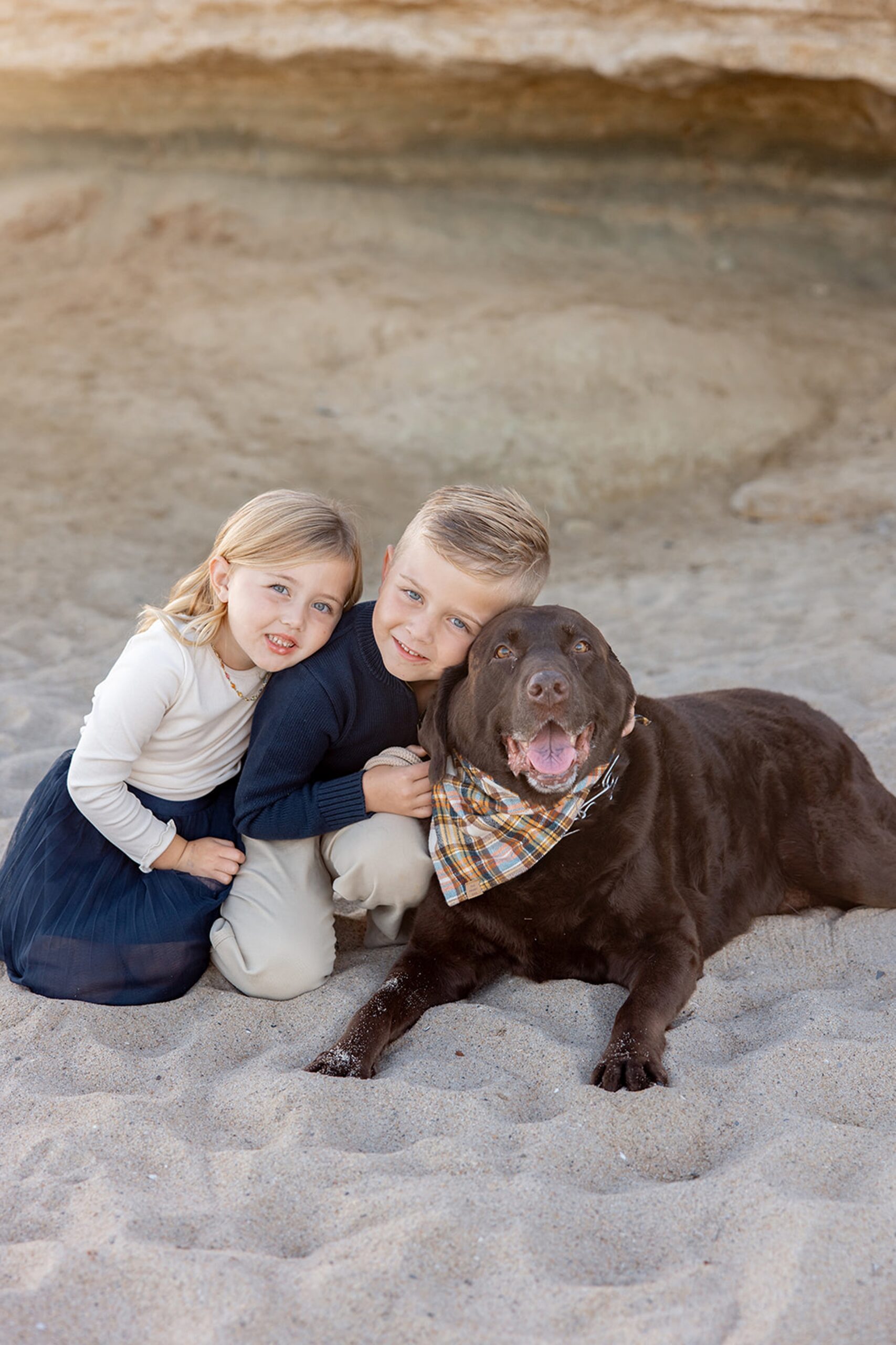 Toddler brother and sister snuggle their brown lab on a beach at sunset after meeting nannies in San Diego