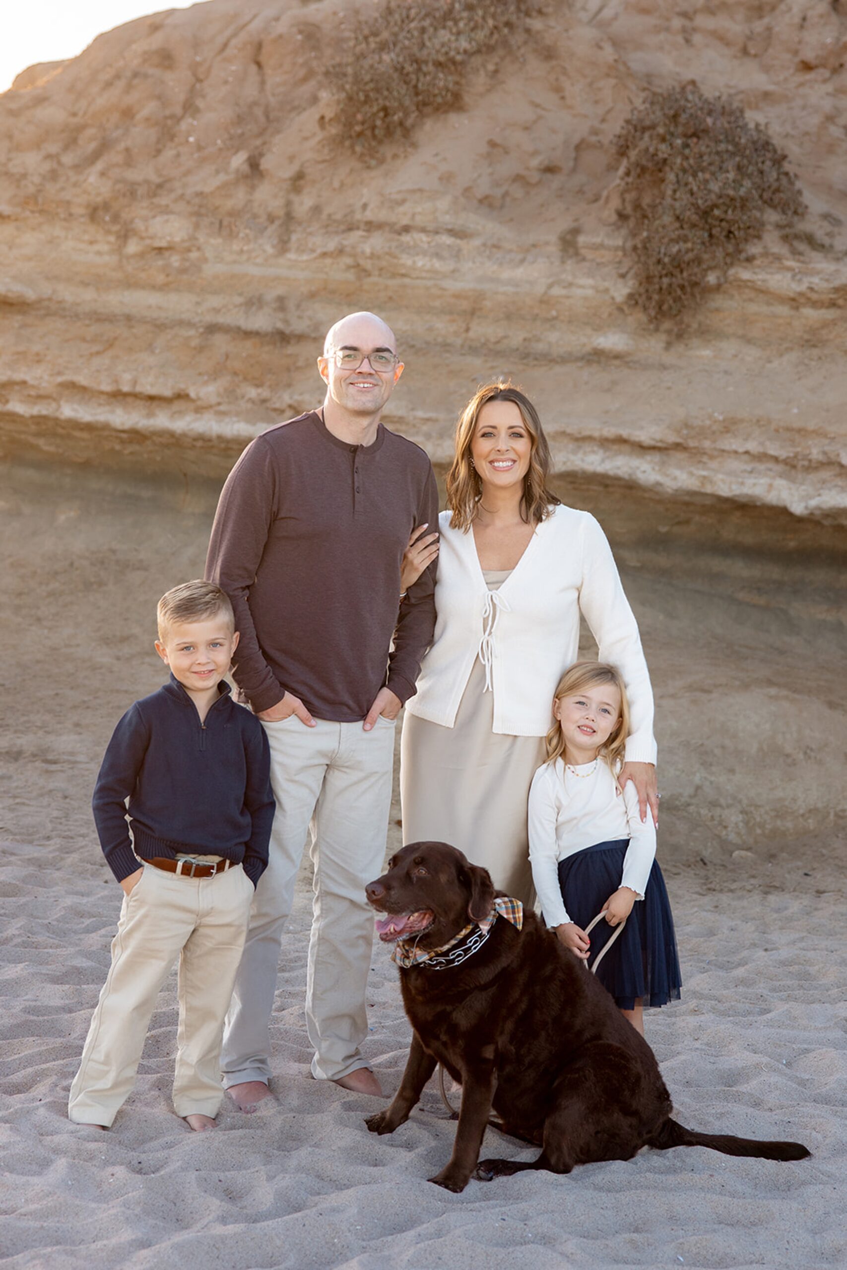 A mom and dad stand on a beach at sunset with their toddler son and daughter and brown lab after finding nannies in San Diego