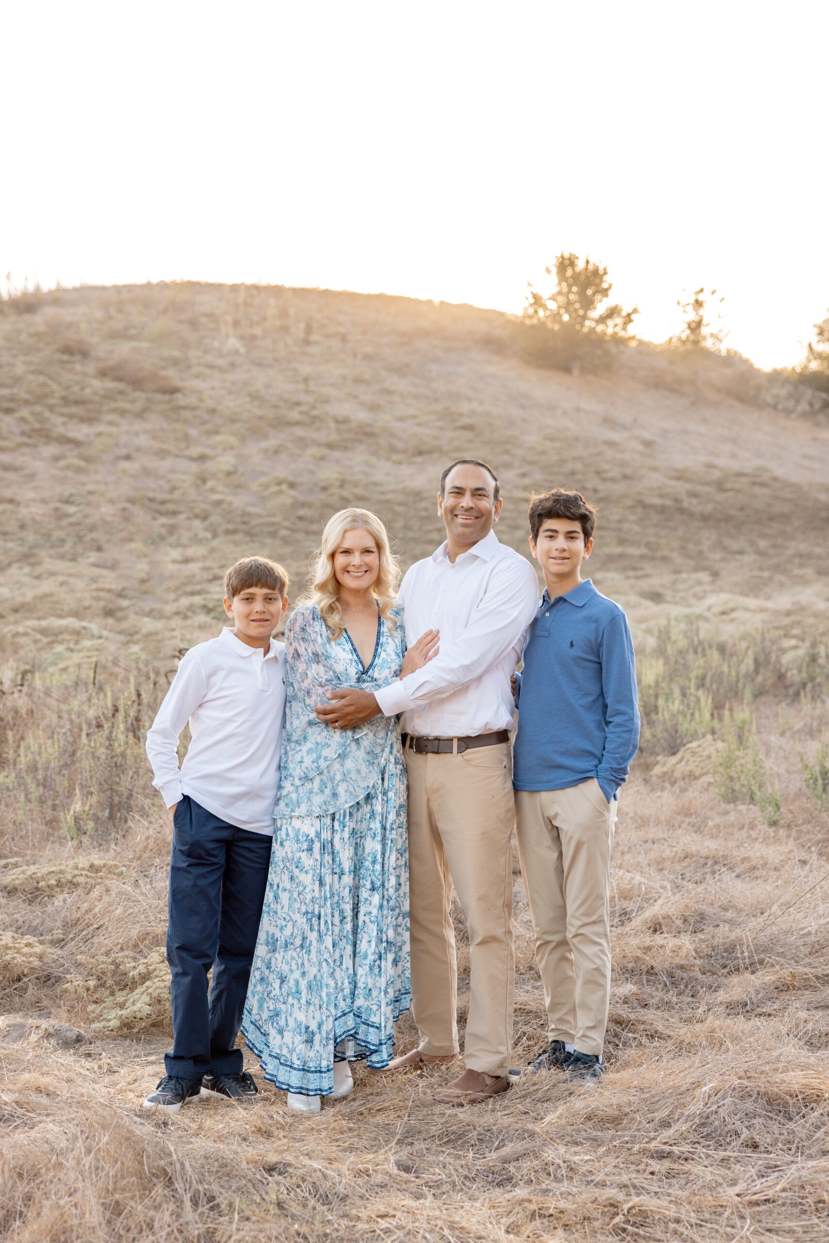 A mom and dad smile together with their two teen suns on a hill at sunset