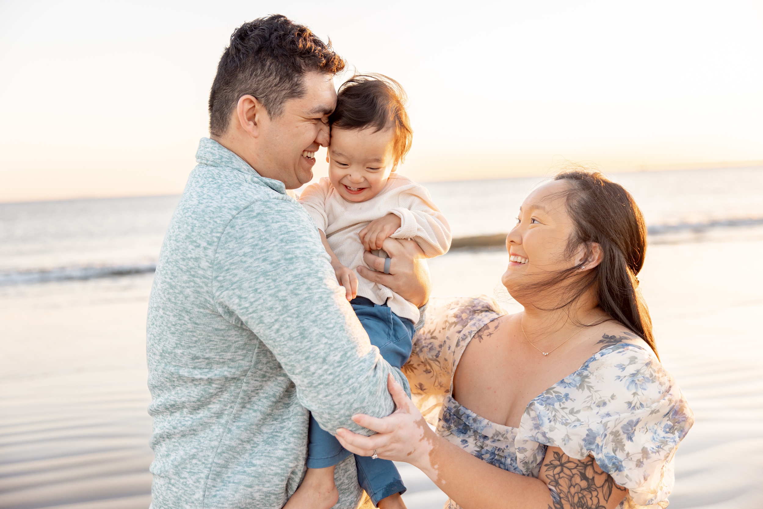 A happy mom and dad snuggle their baby boy on a beach at sunset while all laugh