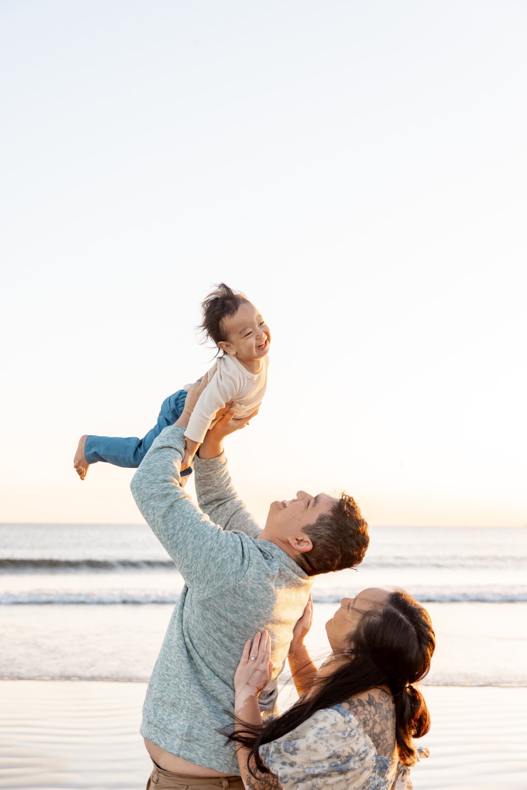 A dad tosses his toddler son in the air on a beach at sunset with mom behind him after visiting toy stores in San Diego