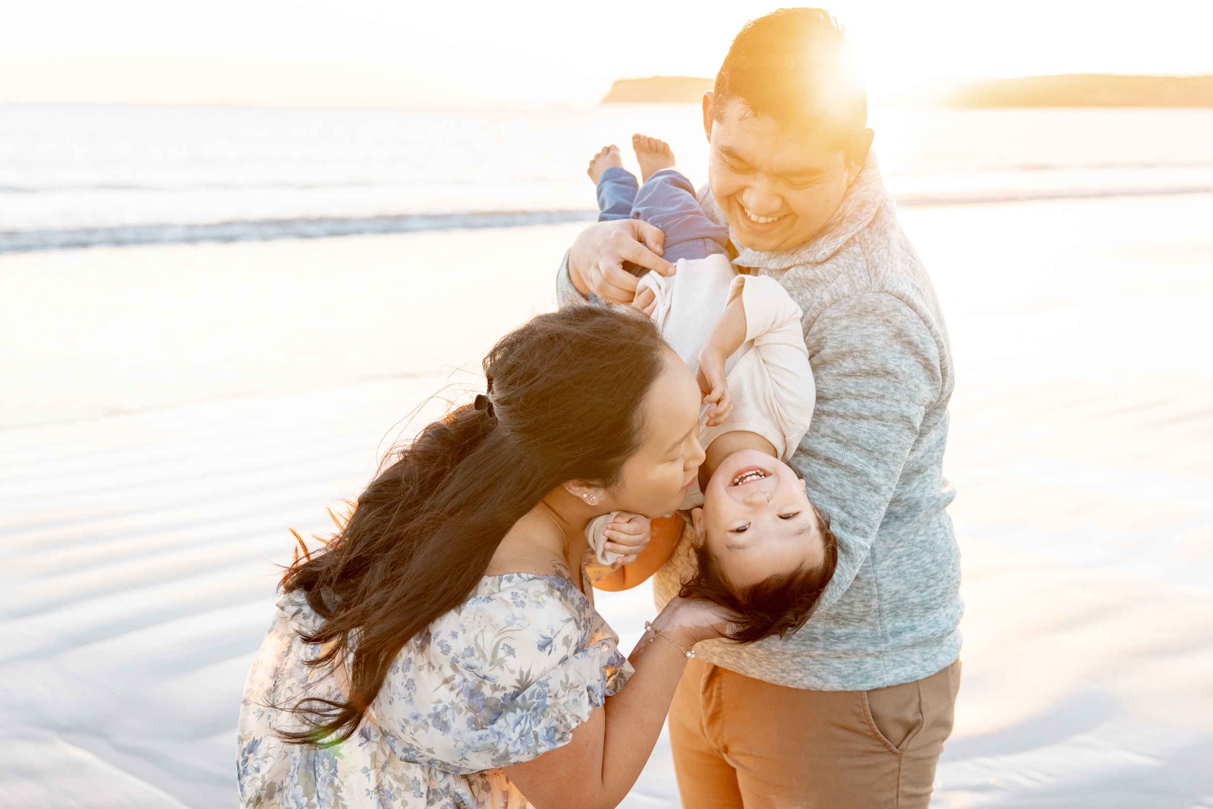 A mom and dad flip and tickle their giggling son on a beach at sunset after visiting toy stores in San Diego