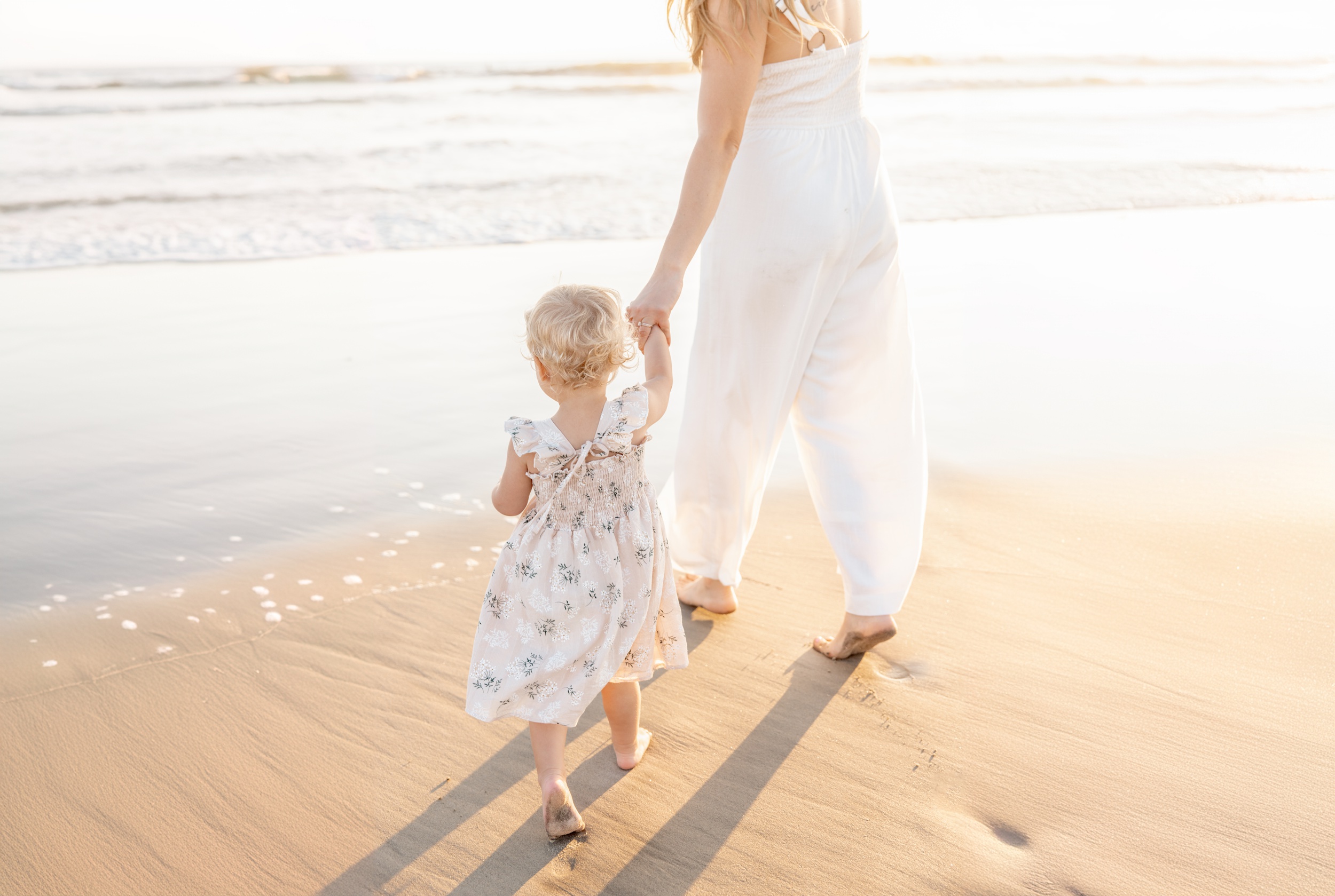 A young girl walks on a beach holding mom's hand at sunset