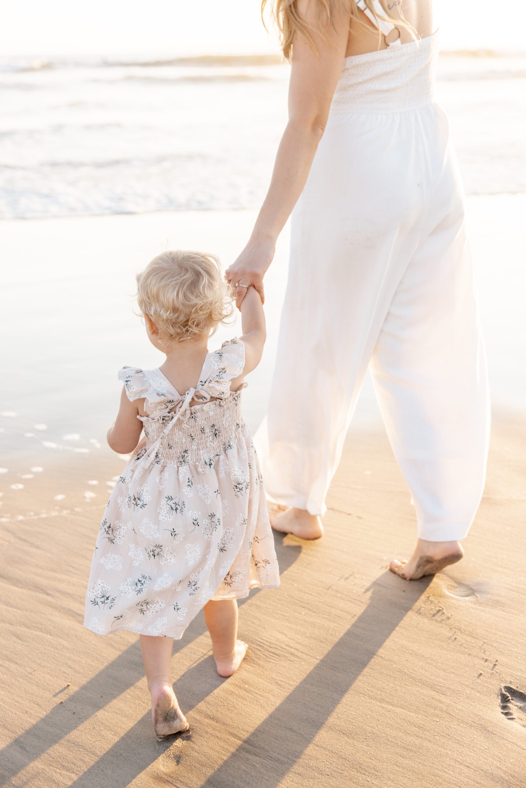 A mom in a white romper walks on a beach holding hands with her toddler daughter in a pink dress at sunset after finding a diaper service in san diego