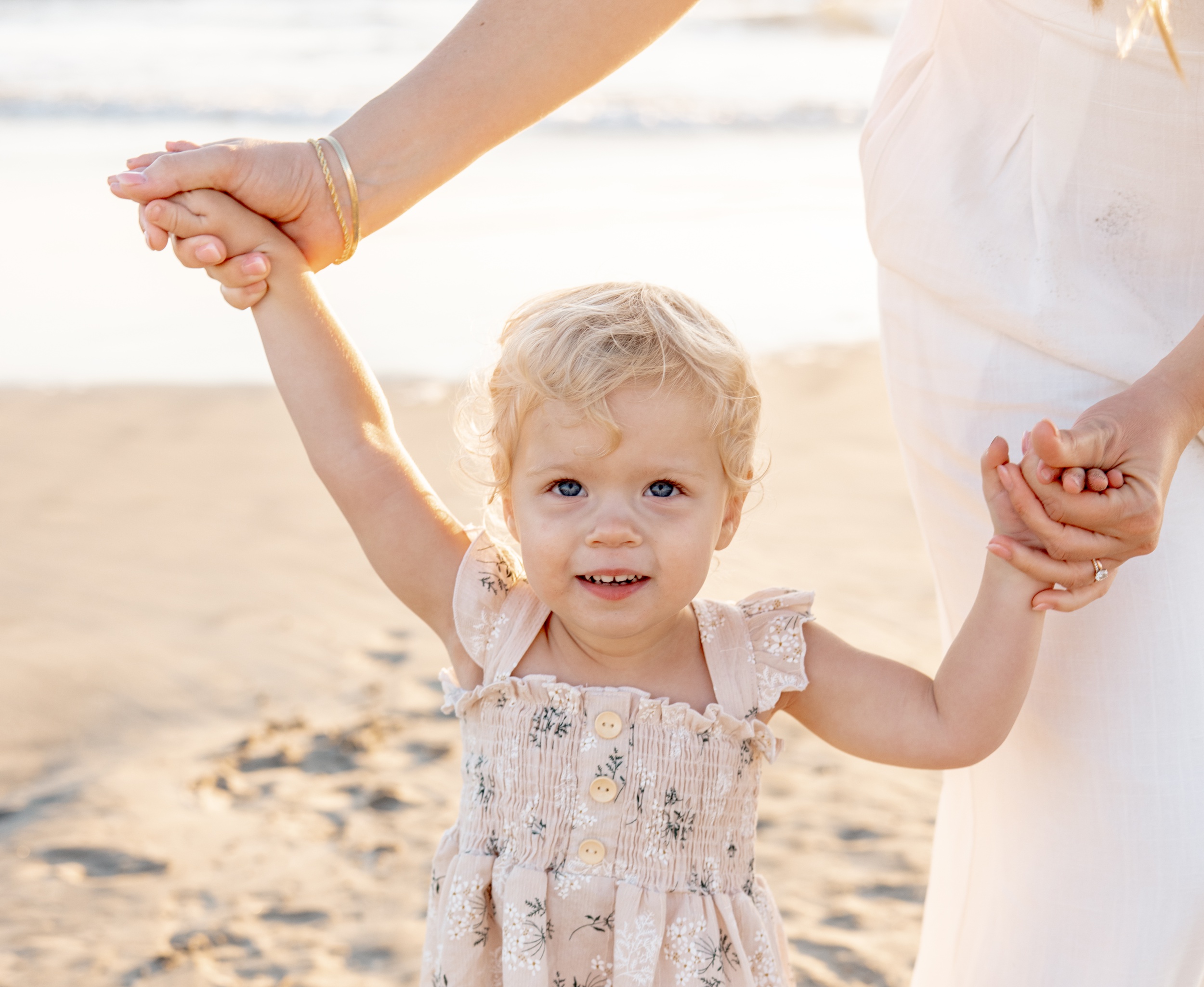 A happy toddler girl in a pink dress walks on a beach with mom's help holding hands after enjoying a diaper service in san diego