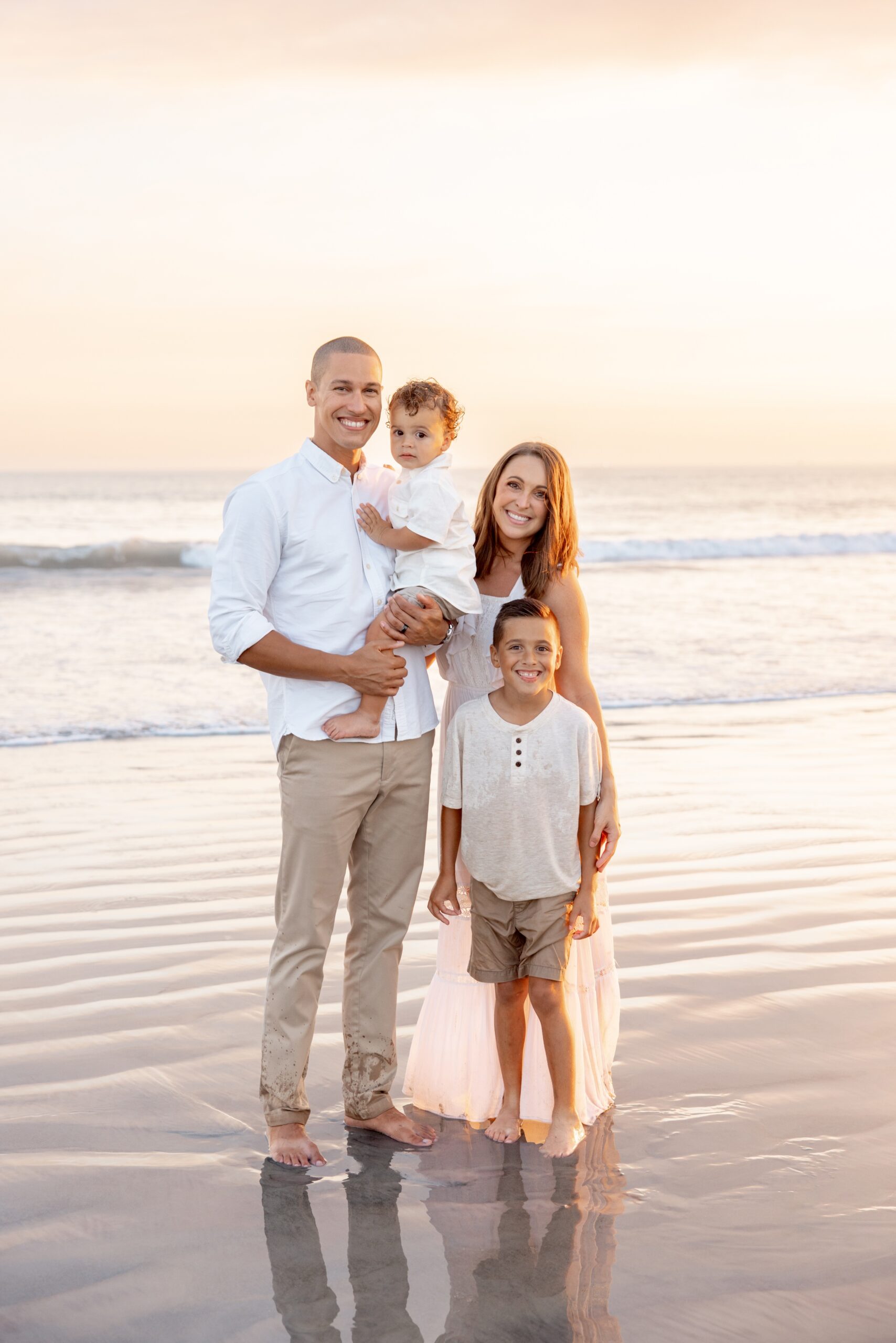 A mom and dad stand smiling on a beach with their toddler in dad's arms and young son standing with mom after some parenting classes in san diego