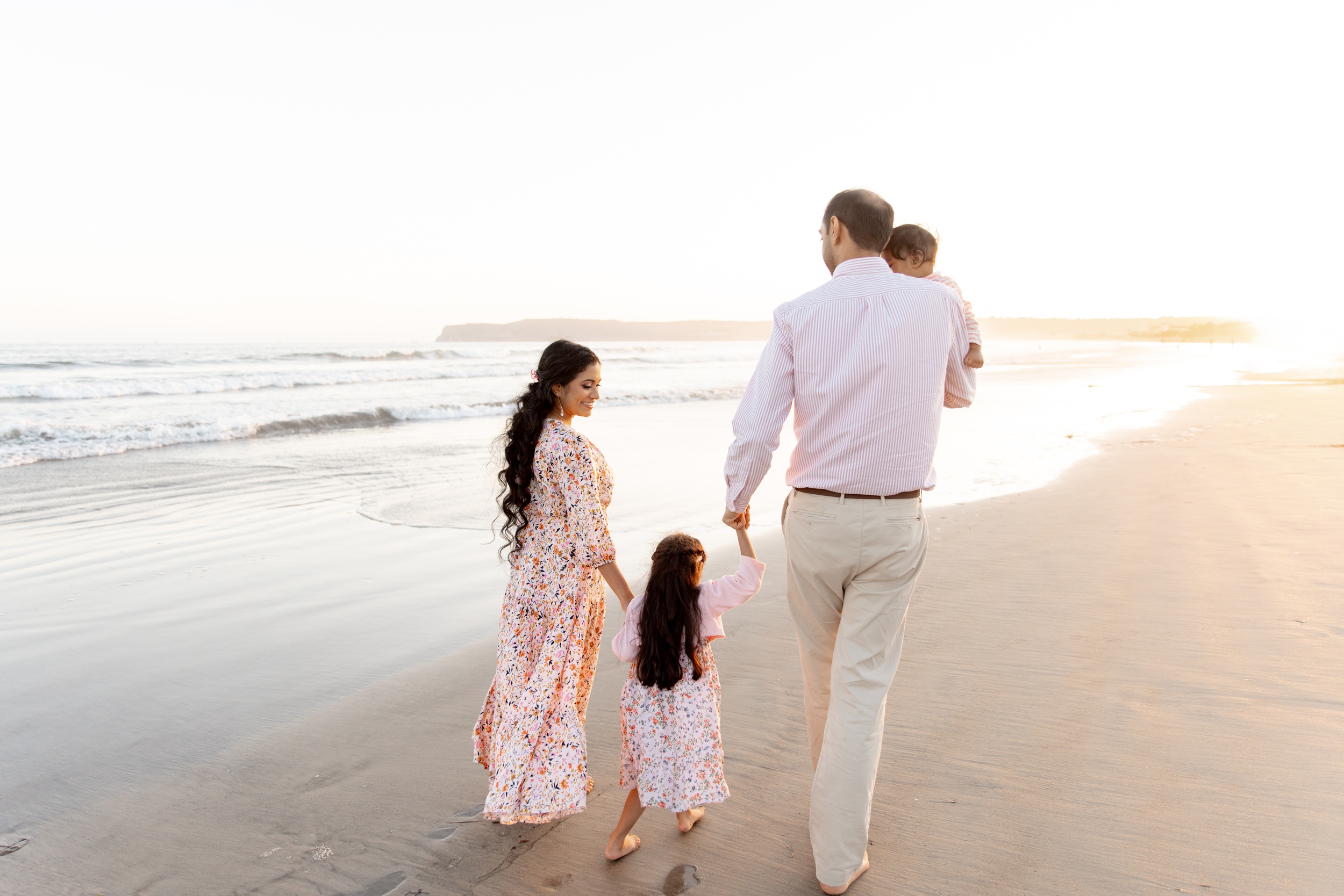 A happy family of 4 with a toddler and baby girls walk on a beach at sunset wearing pink