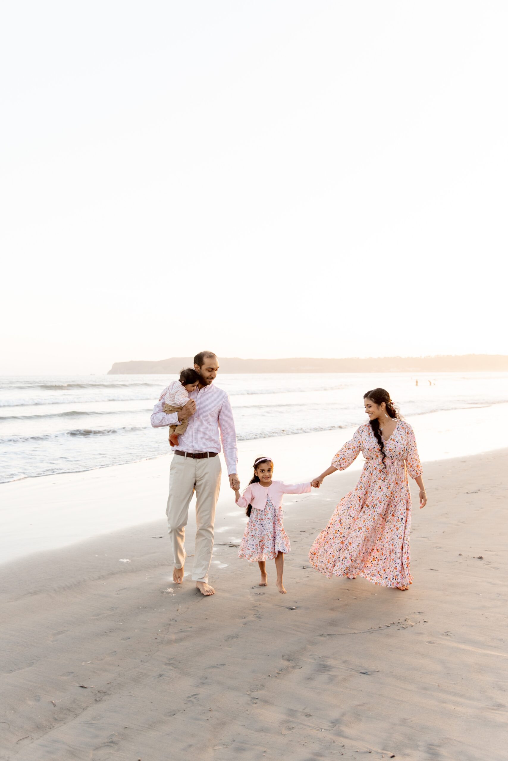 A mom and dad walk on a beach in pink with their baby in dad's arms and holding hands with toddler girl after finding private schools in san diego