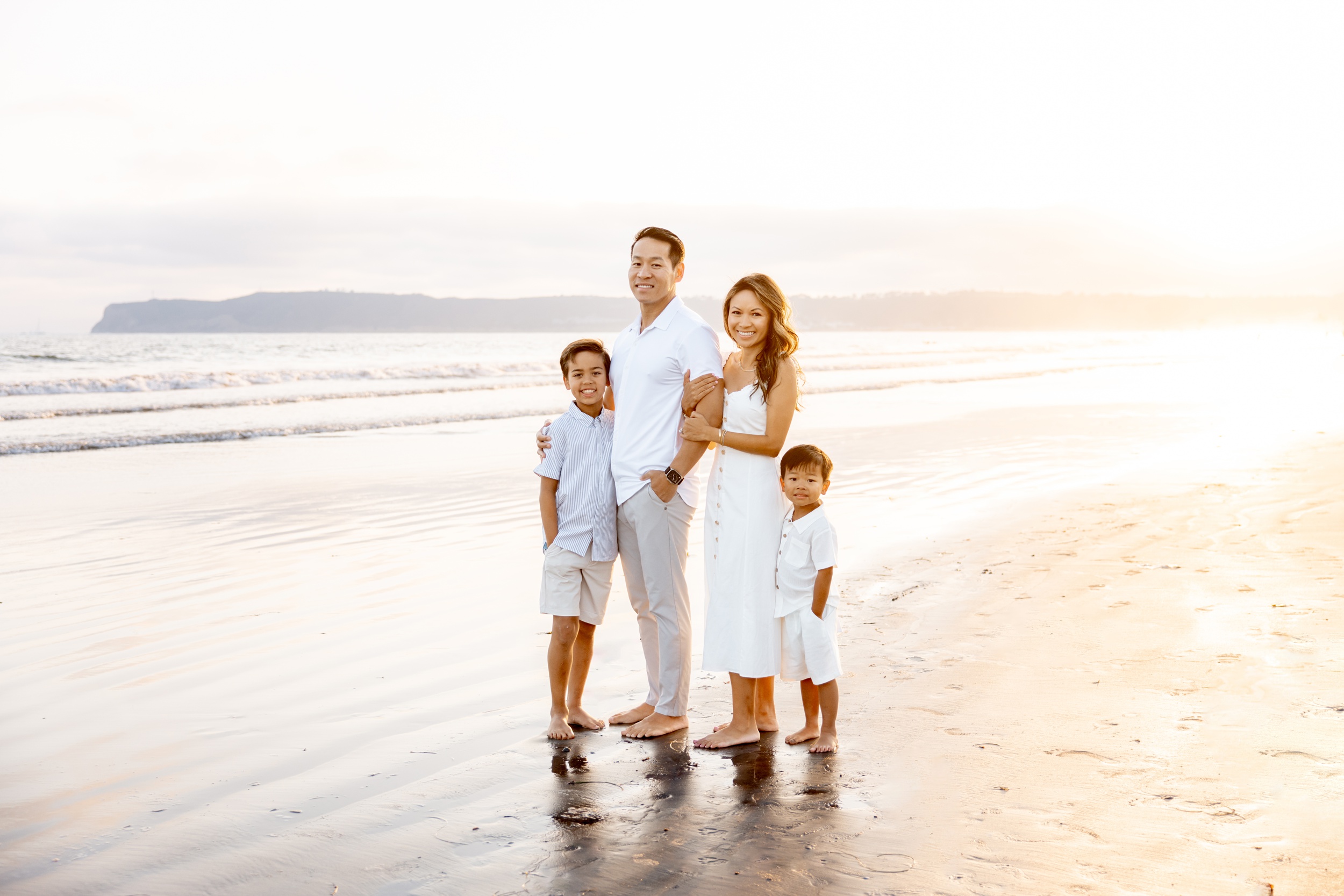 A mom and dad hug while standing on a beach at sunset with their two young sons all in white