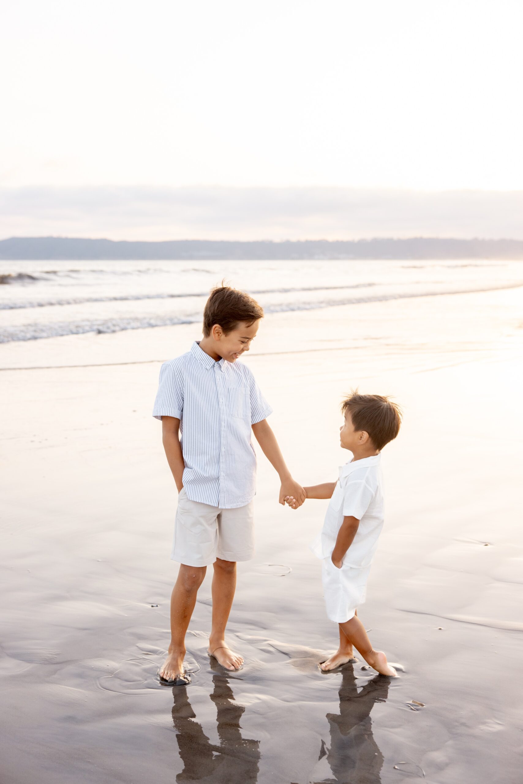 A young boy in white holds hands and laughs with his baby brother on a beach at sunset after visiting splash pads in san diego