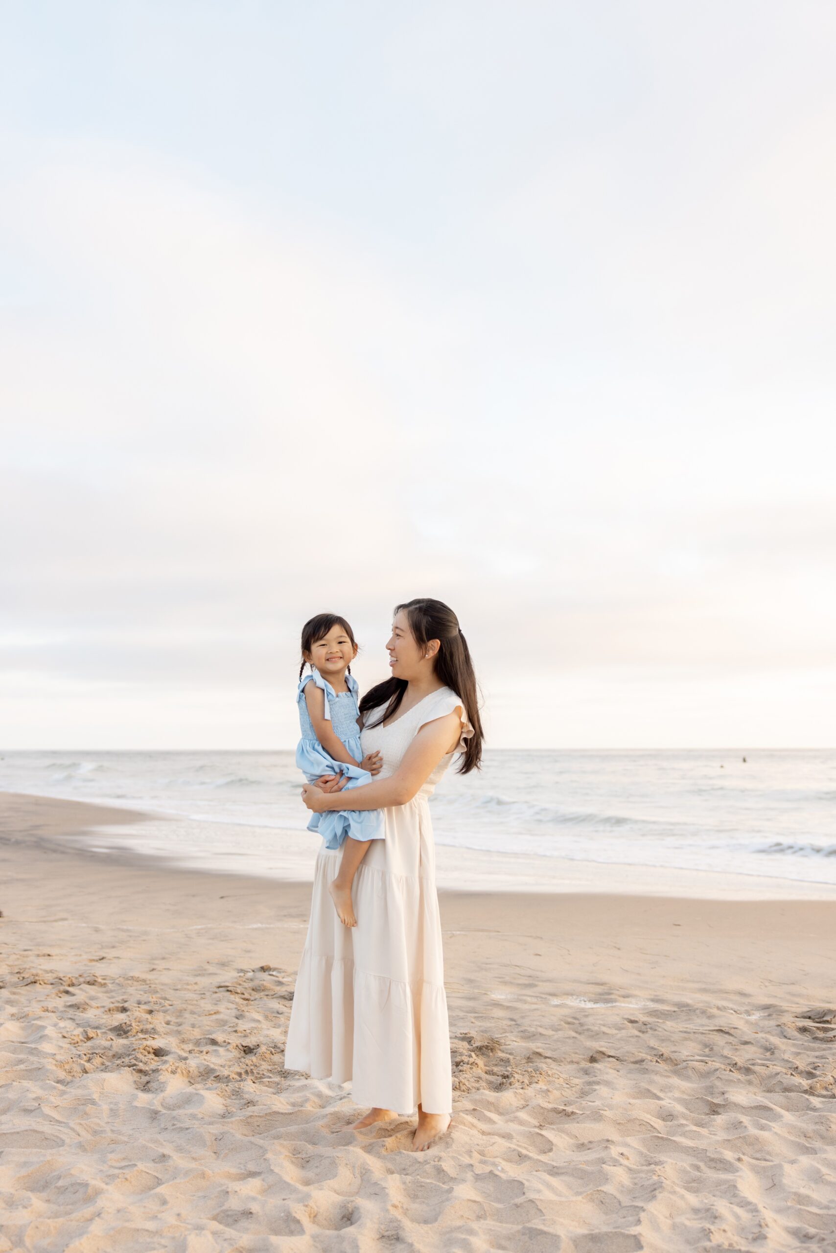 A smiling toddler girl in a blue dress sits in mom's arms on a beach