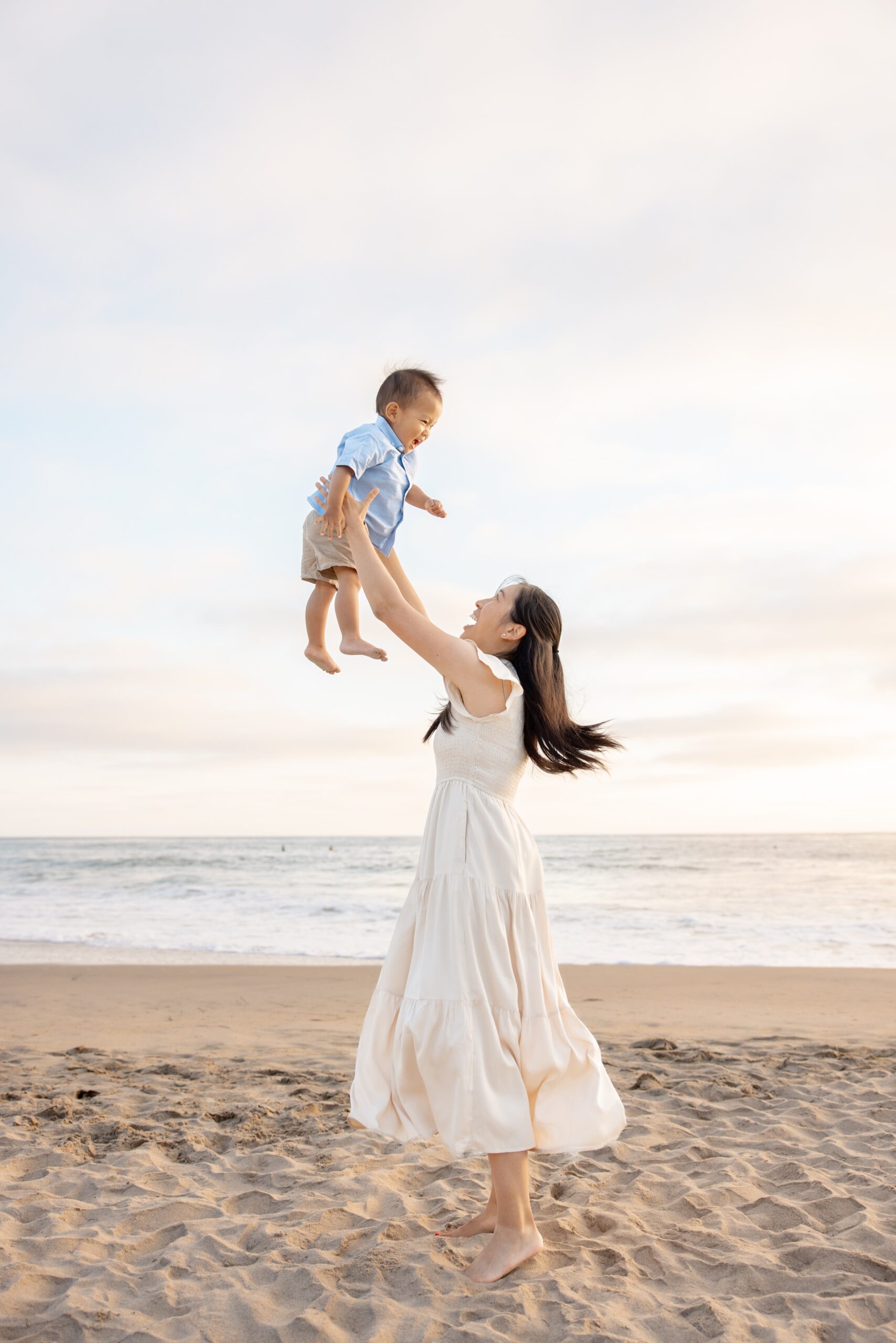 A happy mom in a white dress tosses her baby in the air on a beach at sunset after finding stem summer camps in san diego