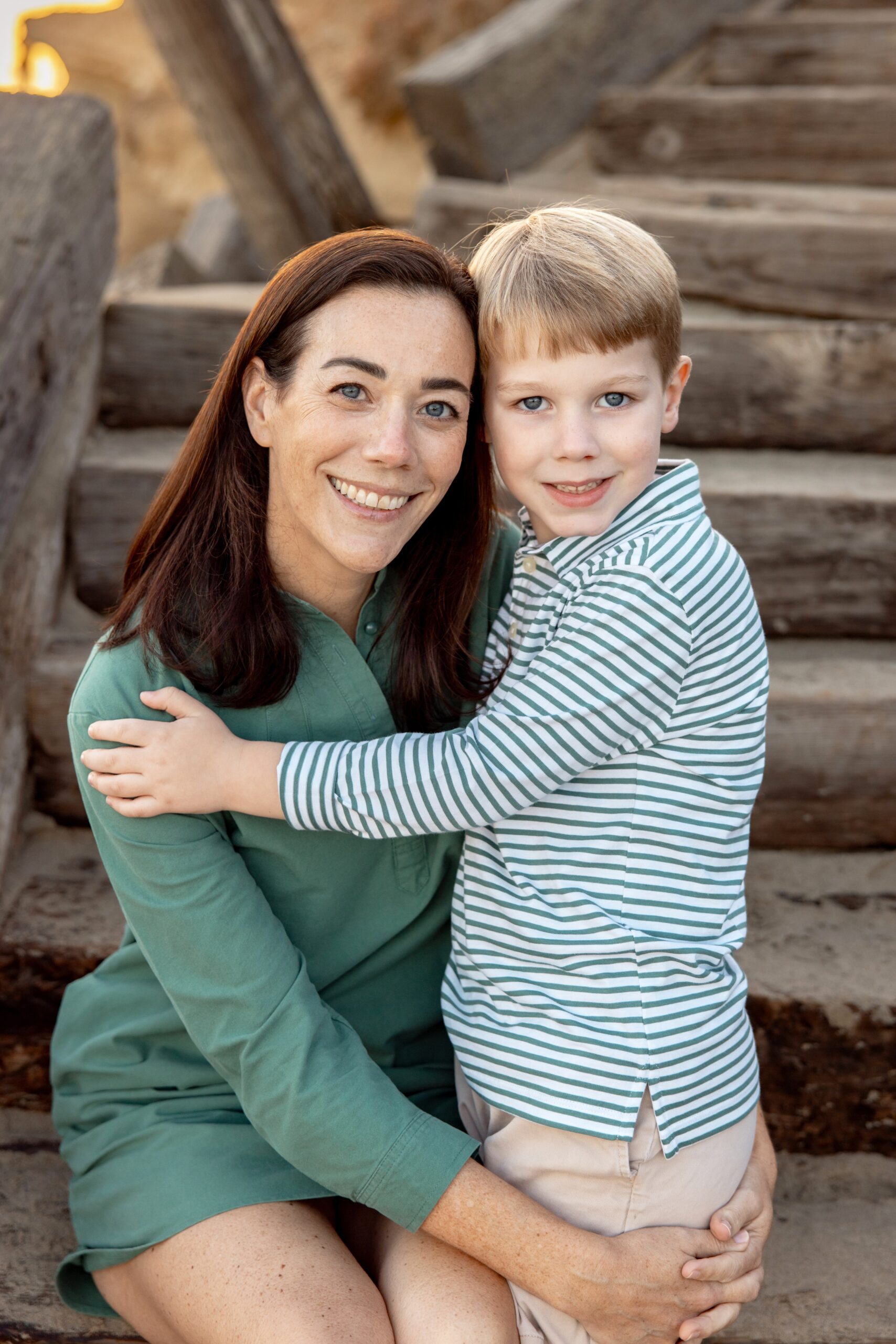 A happy toddler in a stripe shirt hugs mom while sitting on wooden beach stairs at sunset