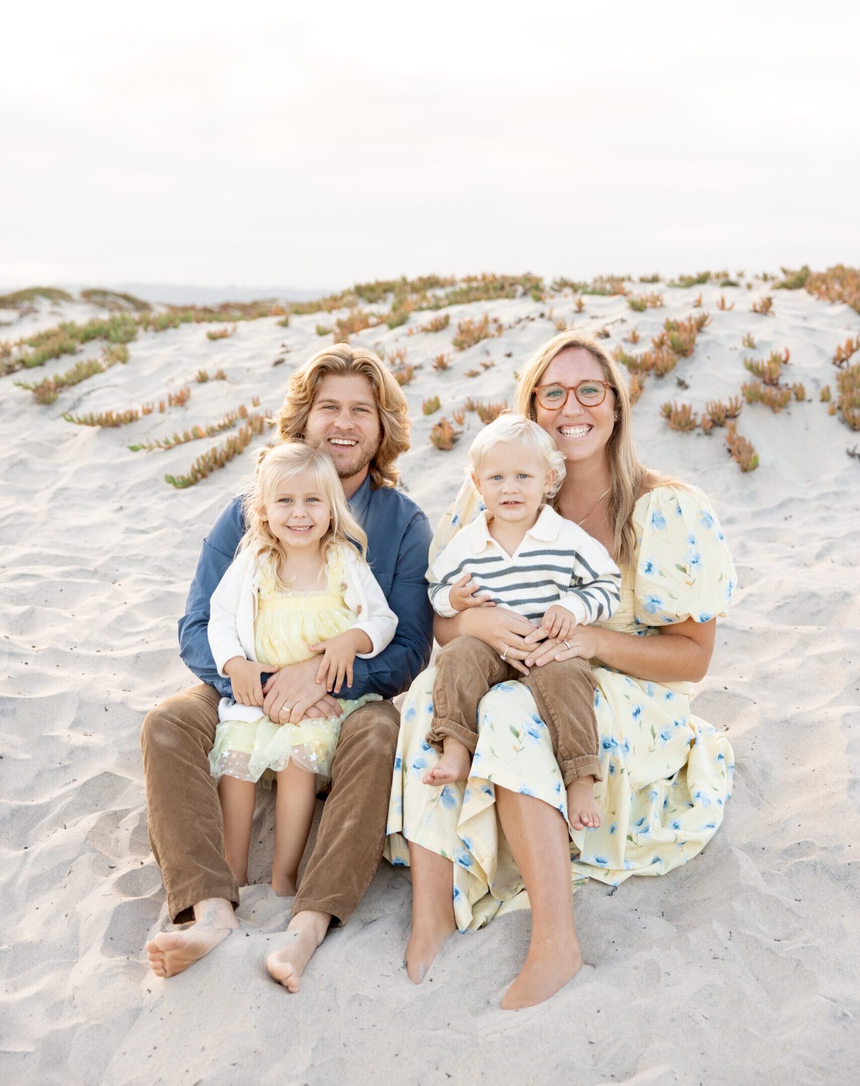 Smiling mom and dad sit on a beach with their toddler son and daughter in their laps while staying at beach resorts in san diego for families