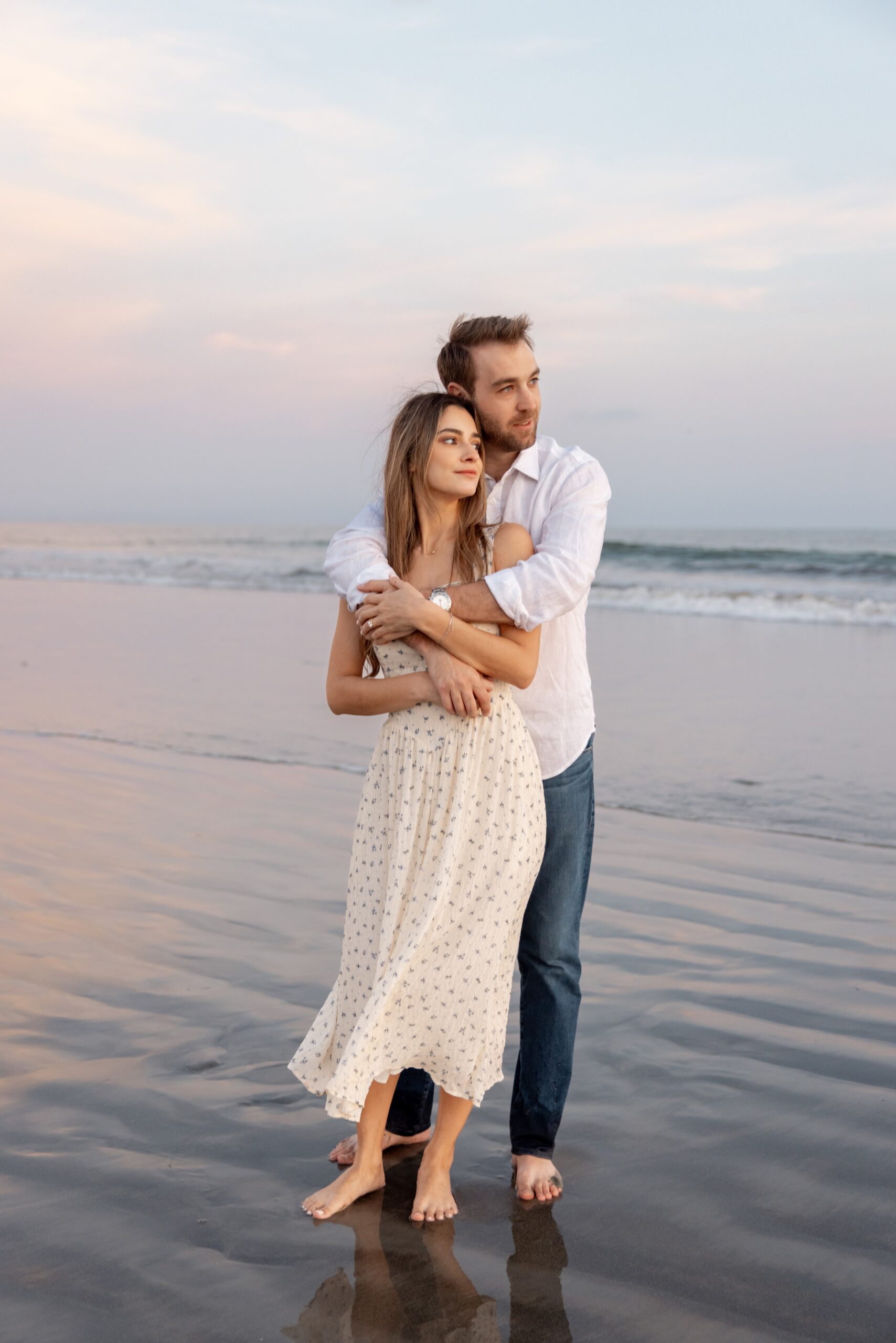 A happy engaged couple snuggles on a beach in the shallows while watching the sunset before their destination wedding in san diego