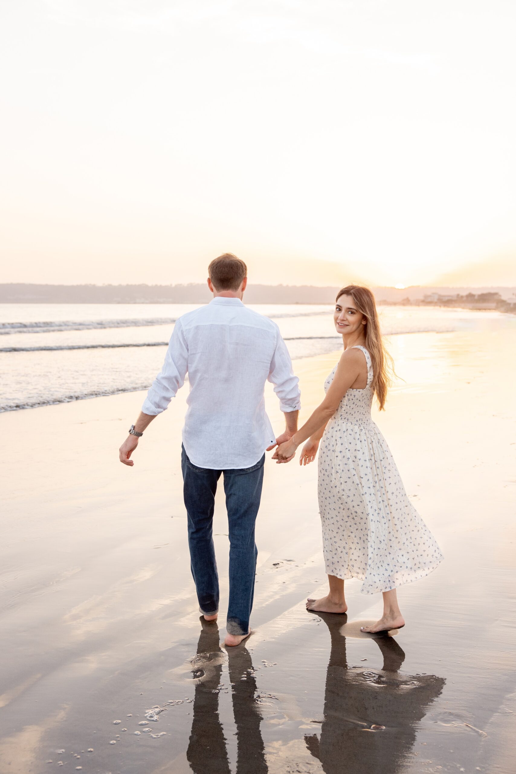 A happy engaged couple in white walk on a beach holding hands at sunset as the woman smiles over her shoulder before their destination wedding in san diego