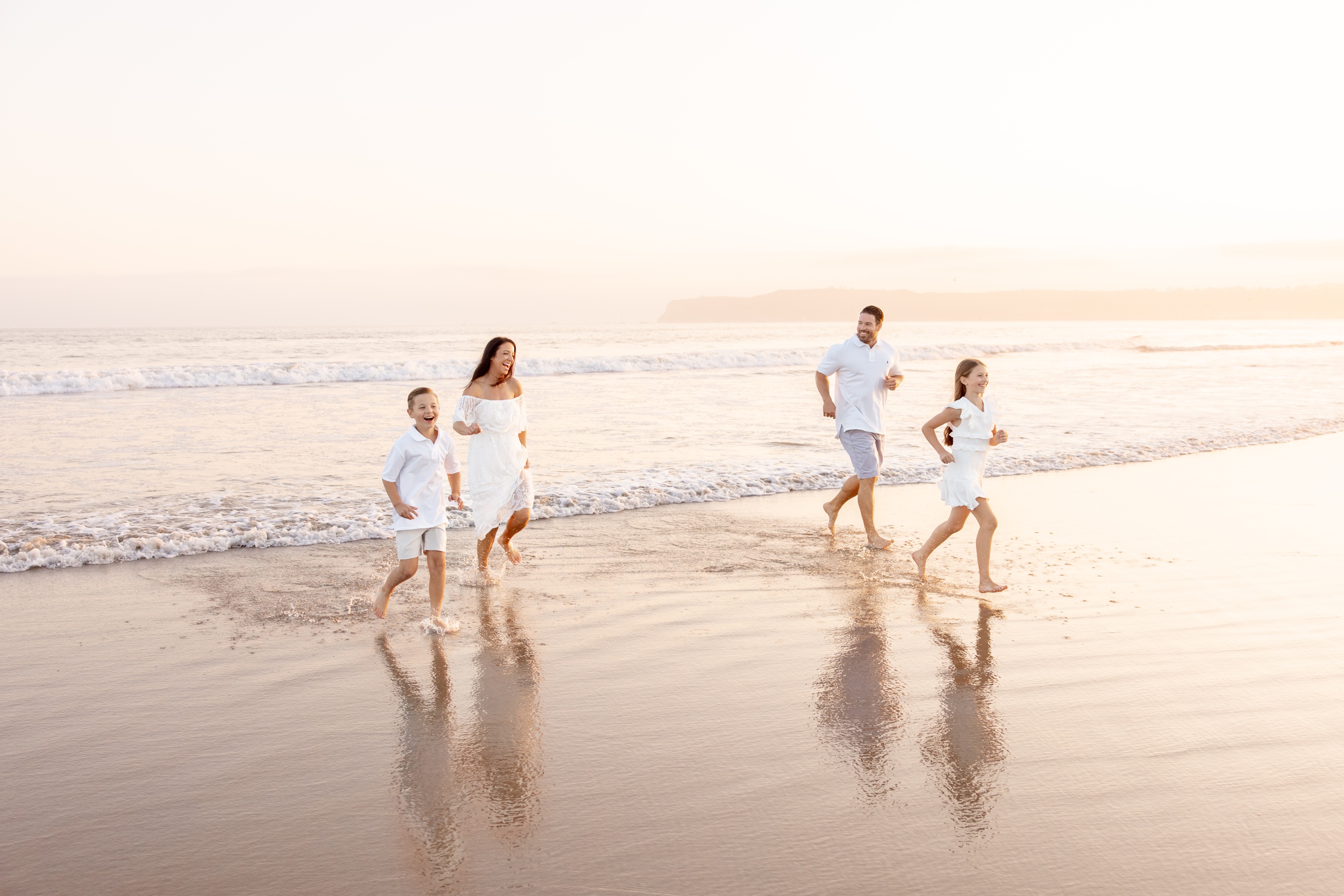 A happy family in all white outfits run on a beach at sunset while laughing