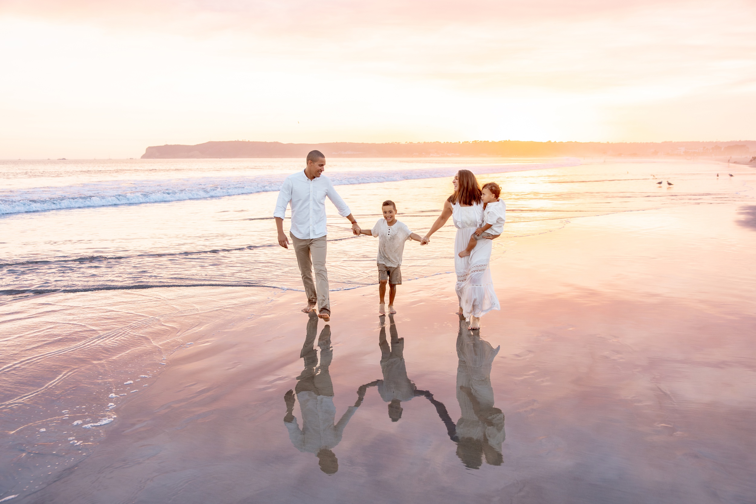 A mom and dad laugh while walking holding hands on a beach with their toddler son and one on mom's hip