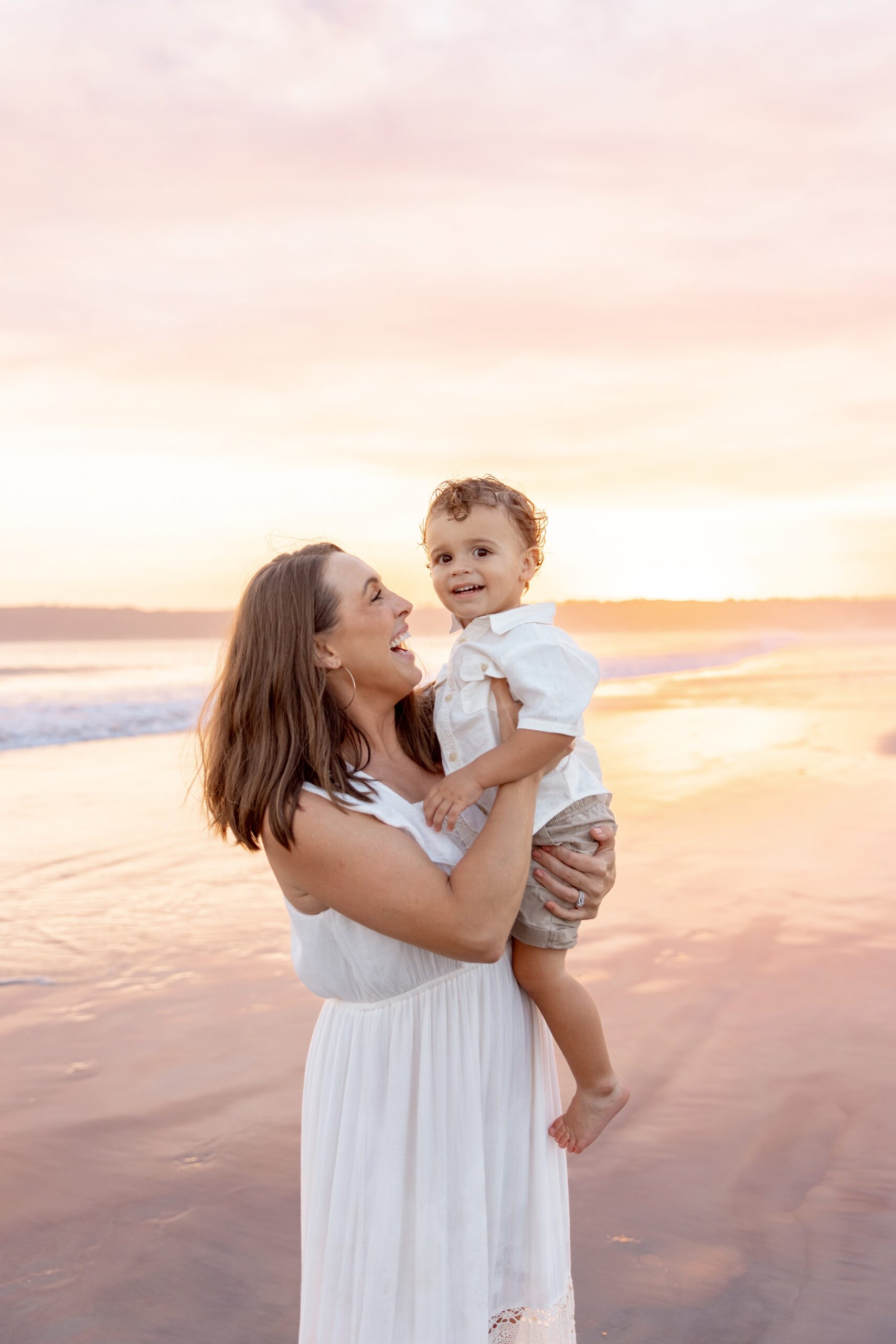 A happy mom in a white dress holds and tickles her toddler son on a beach before visiting restaurants with play areas in san diego